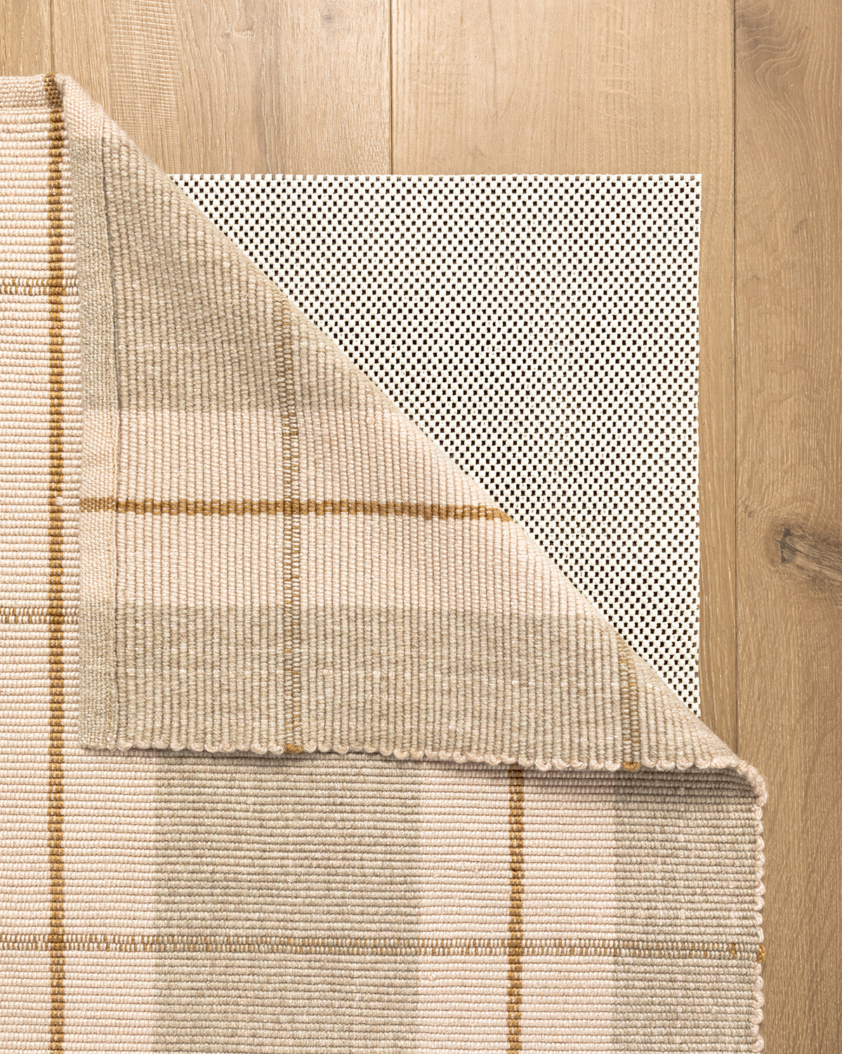A beige and tan plaid area rug is partially folded to show the Premium Outdoor Rug Pad, featuring a black and white dotted design, on a light wood floor, by McGee & Co.