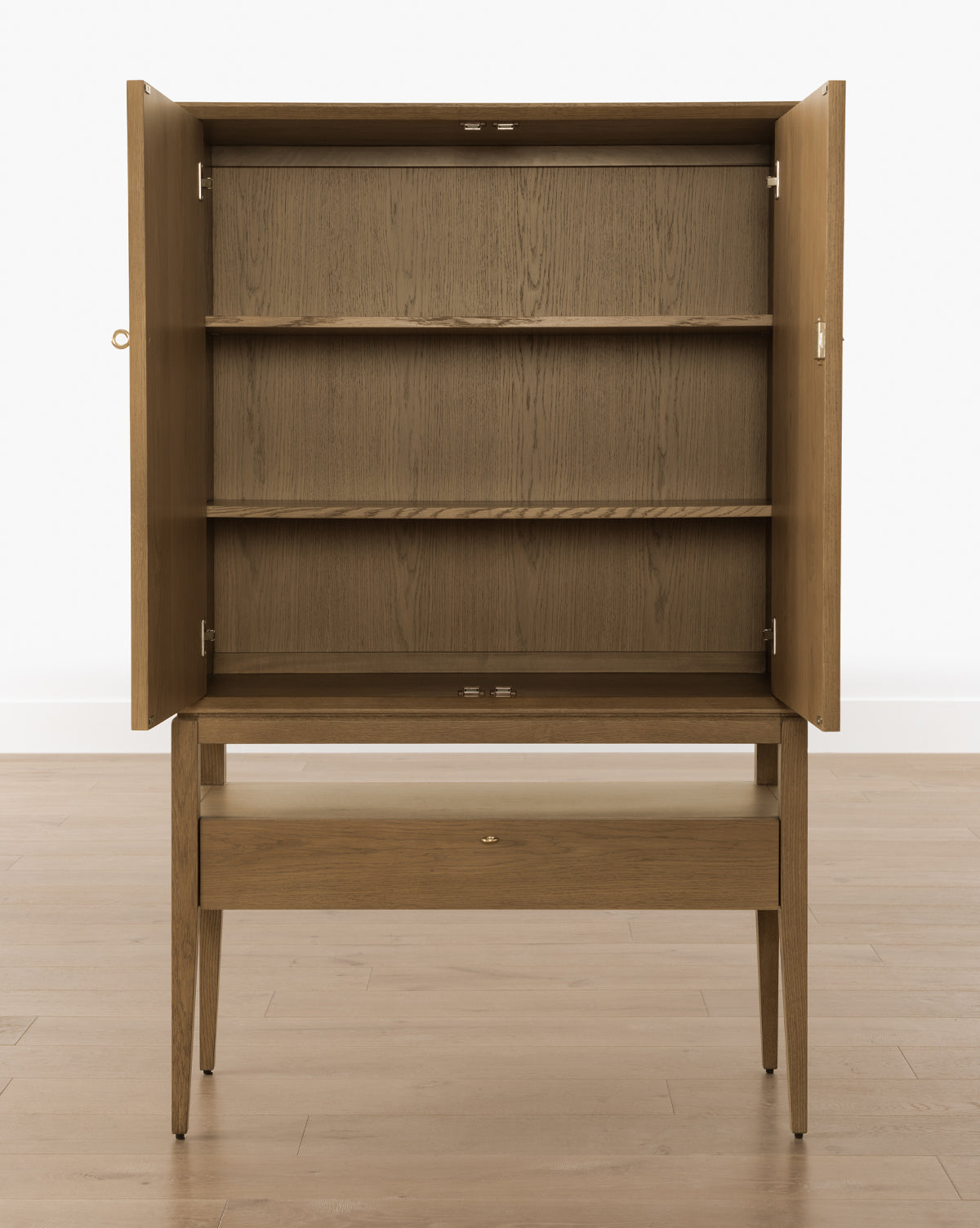 The Jahnsen Cabinet features a honey oak finish, two open doors revealing shelves, a closed bottom drawer, and stands on four tapered legs, by McGee & Co.
