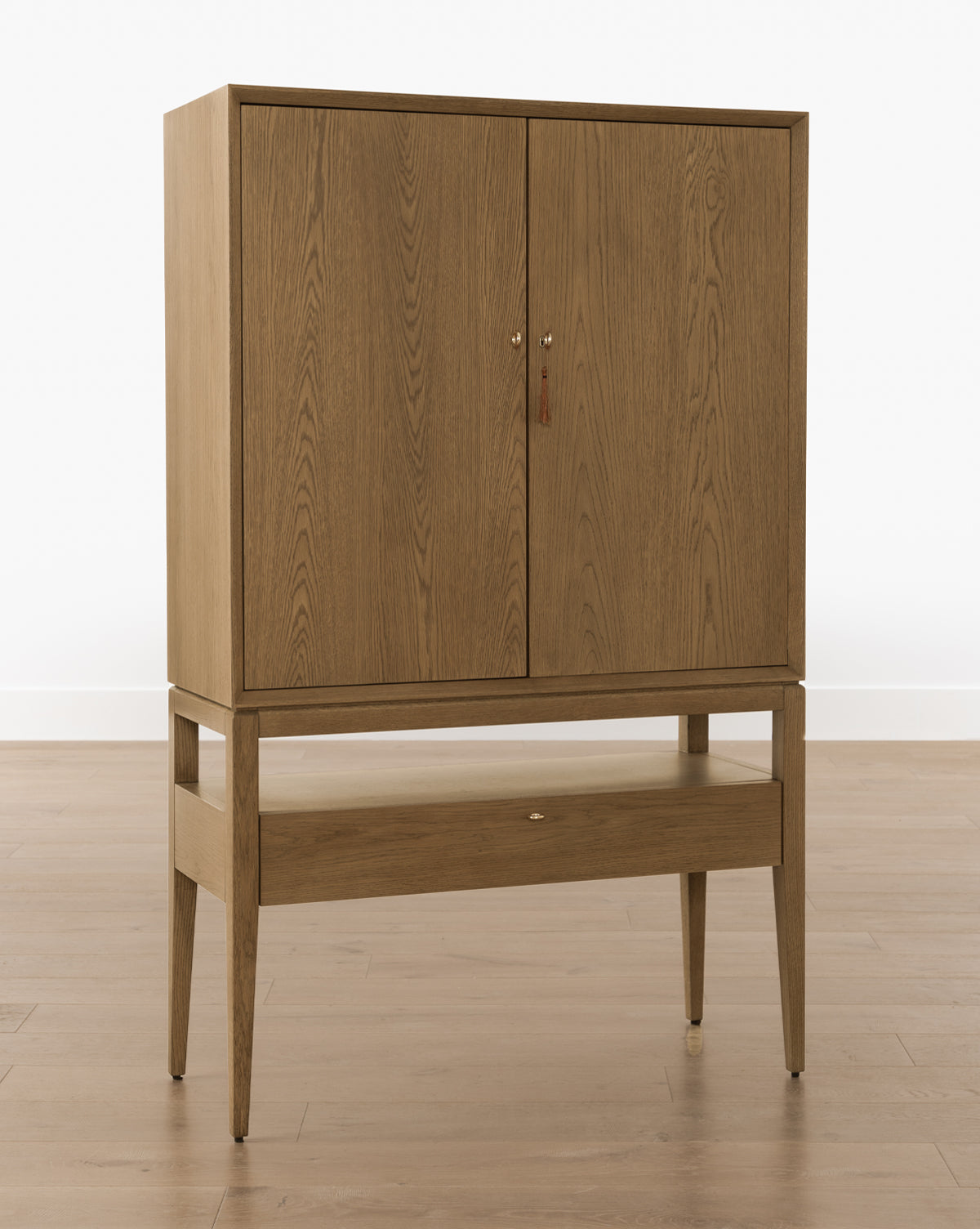 The Jahnsen Cabinet is a mid-century modern piece with a honey oak finish, two upper doors, a lower drawer, and four tapered legs, displayed on a light wood floor against a white wall, by McGee & Co.