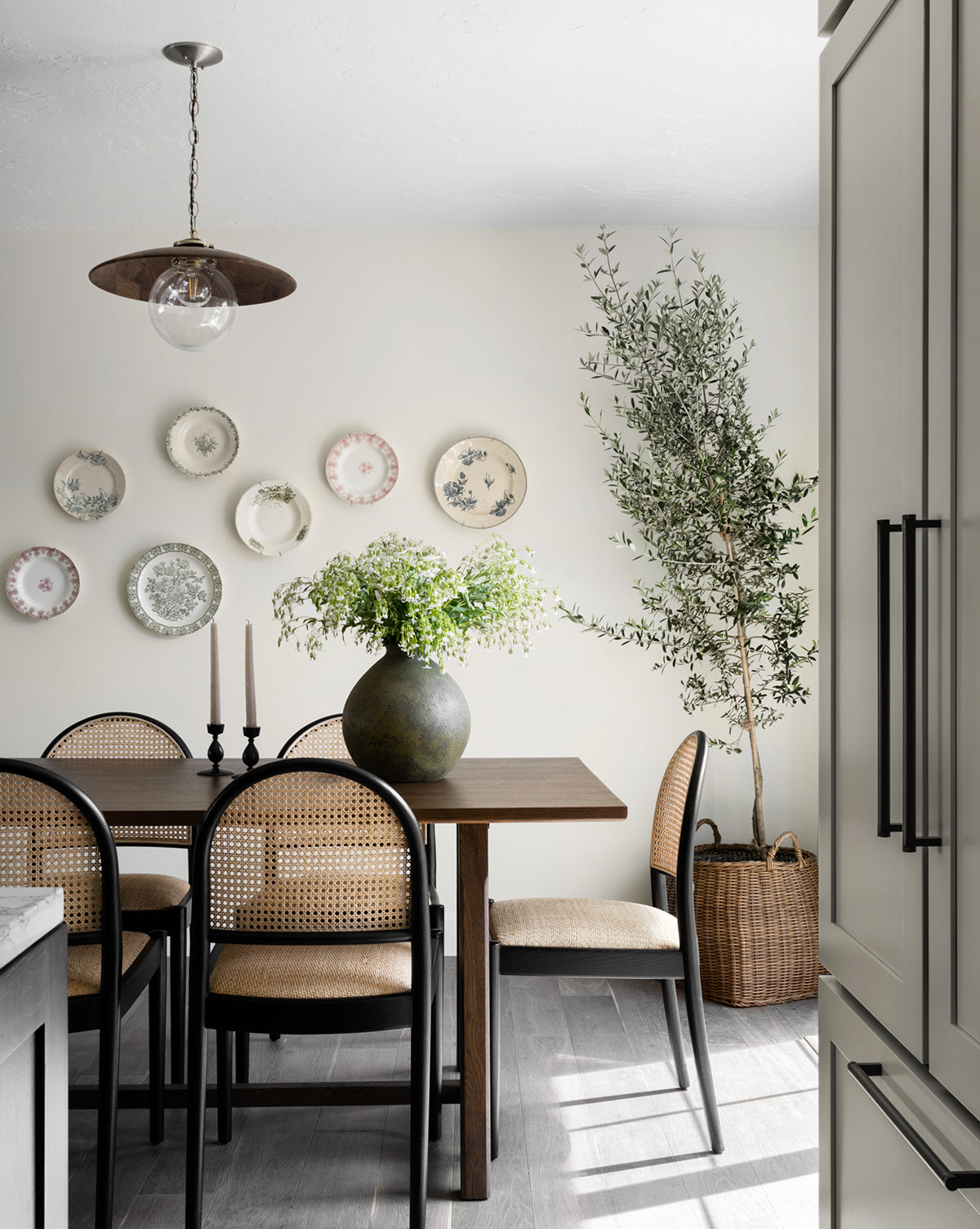 A dining room with a wooden table, four cane-back chairs, a large vase with greenery, two black candles, indoor plants in a Handled Planter Basket, decorative plates on a light wall, and a pendant light overhead.