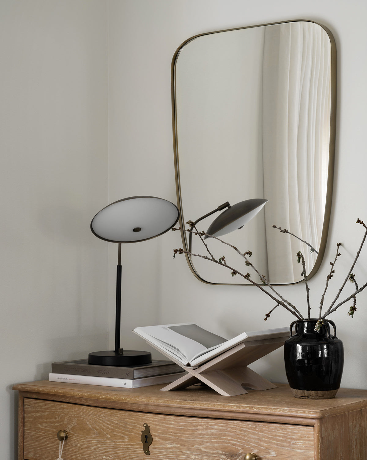 A modern corner features a light wood dresser, black table lamp, open book on stand, black vase with branches, and the Jace Inset Mirror with a sleek stainless-steel frame reflecting airy white curtains by McGee & Co.