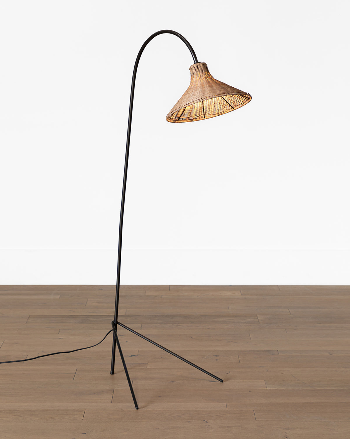 The Johanson Woven Floor Lamp features a black, slender, curved metal stand and a cone-shaped rattan shade, standing on a wooden floor against a plain white wall, by McGee & Co.