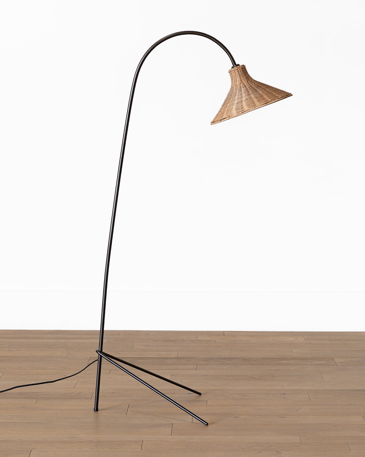 The Johanson Woven Floor Lamp, featuring a curved black metal stand, tripod base, and woven shade, stands on a wooden floor against a plain white wall, by McGee & Co.