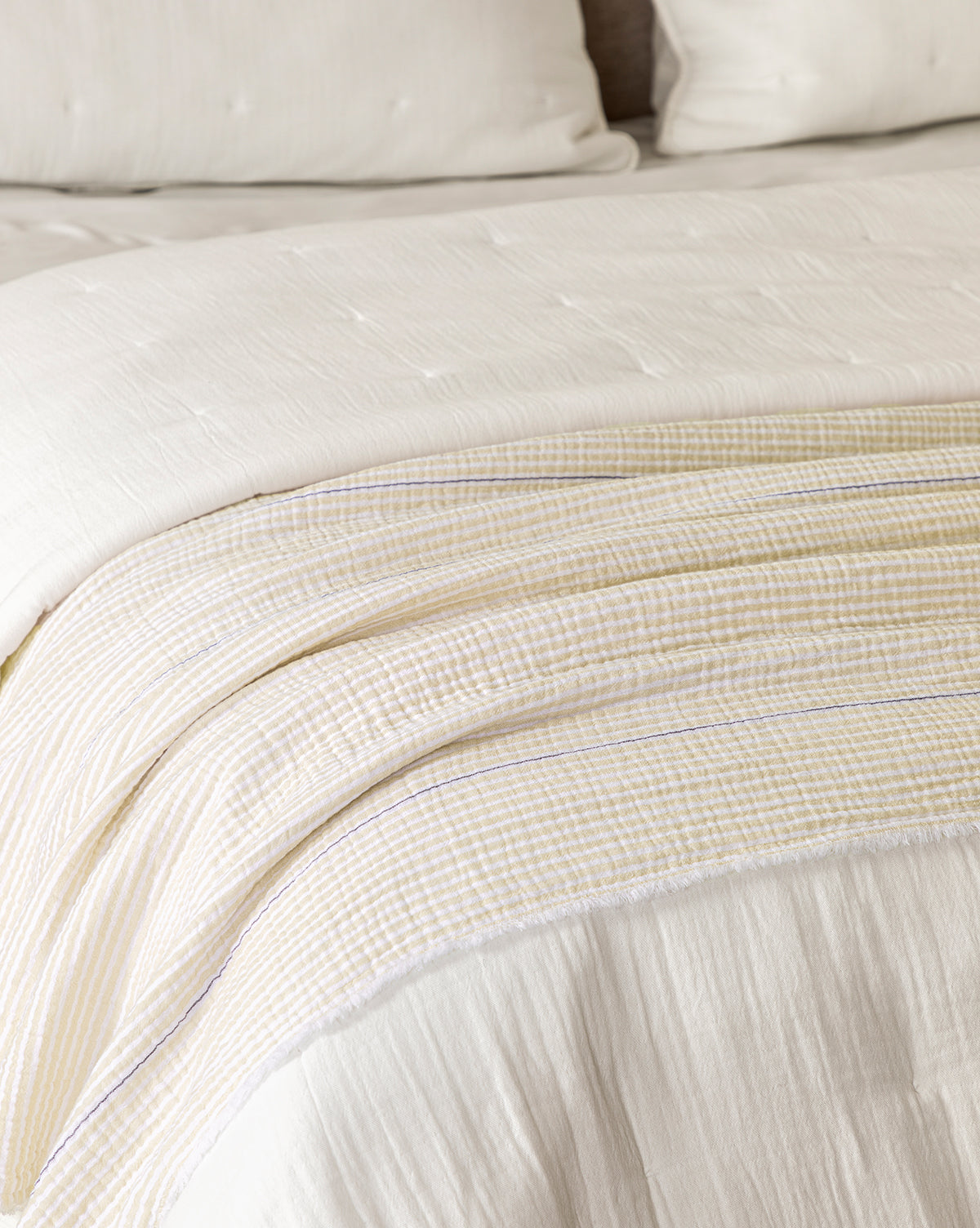 A close-up of a neatly made bed features the Keats Coverlet, a textured white cotton layer. A light, off-white striped throw and two matching pillows enhance the breathable bedding, highlighting comfort and style by McGee & Co.