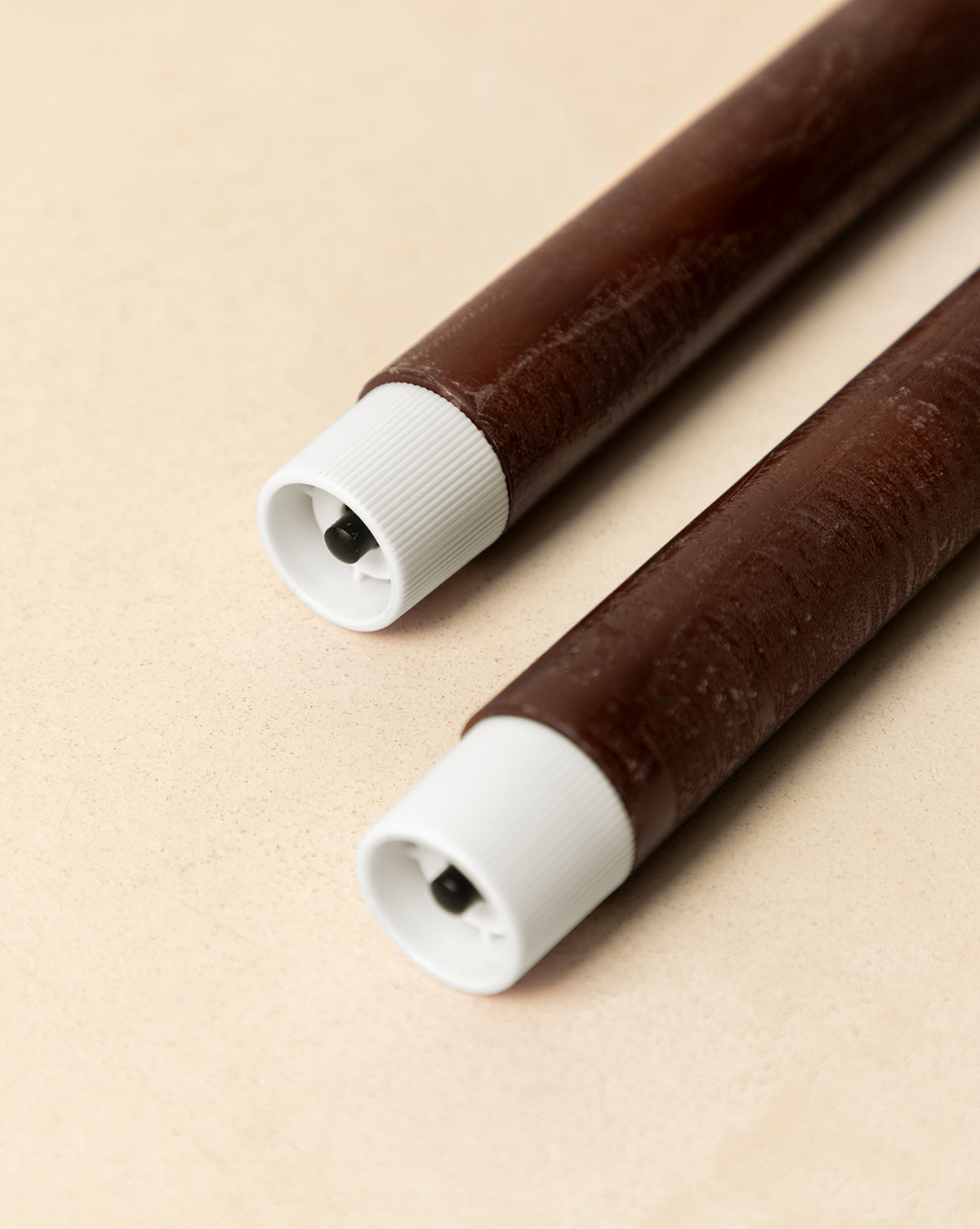 A set of two brown LED Wax Taper Candles with white caps and small black circular openings, lying parallel on a light beige surface, by McGee & Co.