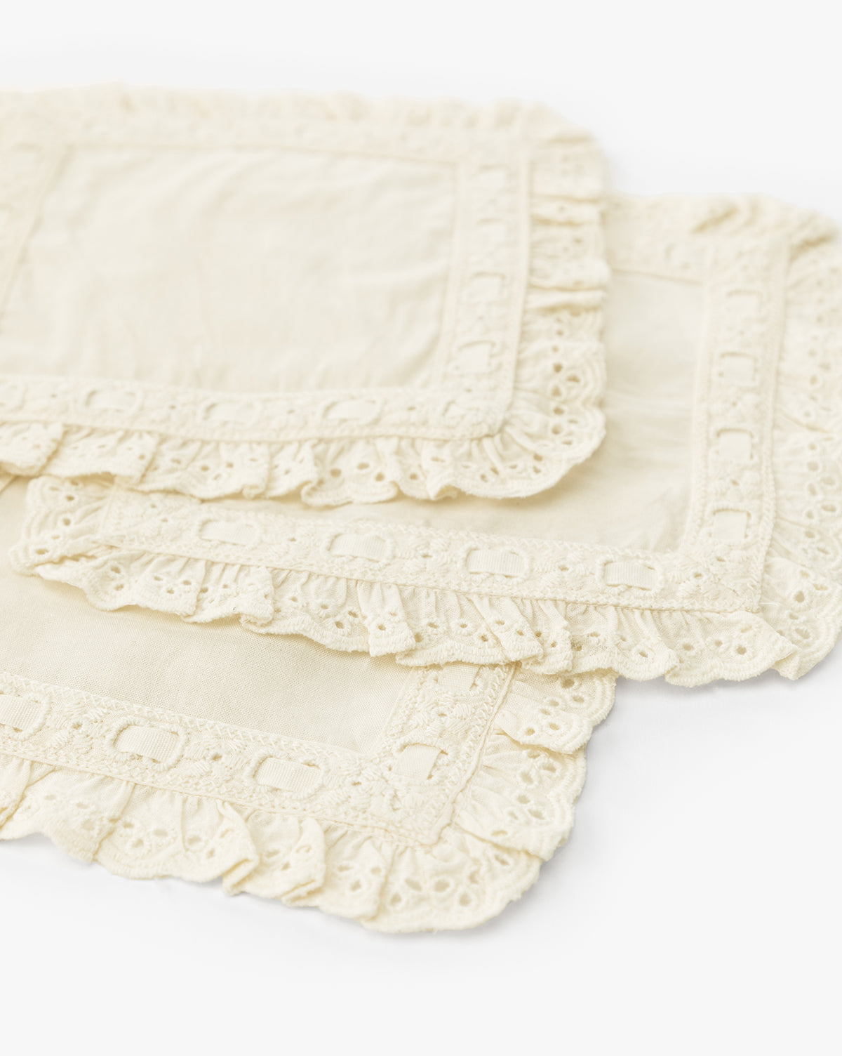 Four cream-colored, square Linen Lace Trim Cocktail Napkins are stacked slightly askew on a white background, featuring intricate eyelet lace borders—ideal as vintage-inspired napkins for elegant table decor, by McGee & Co.