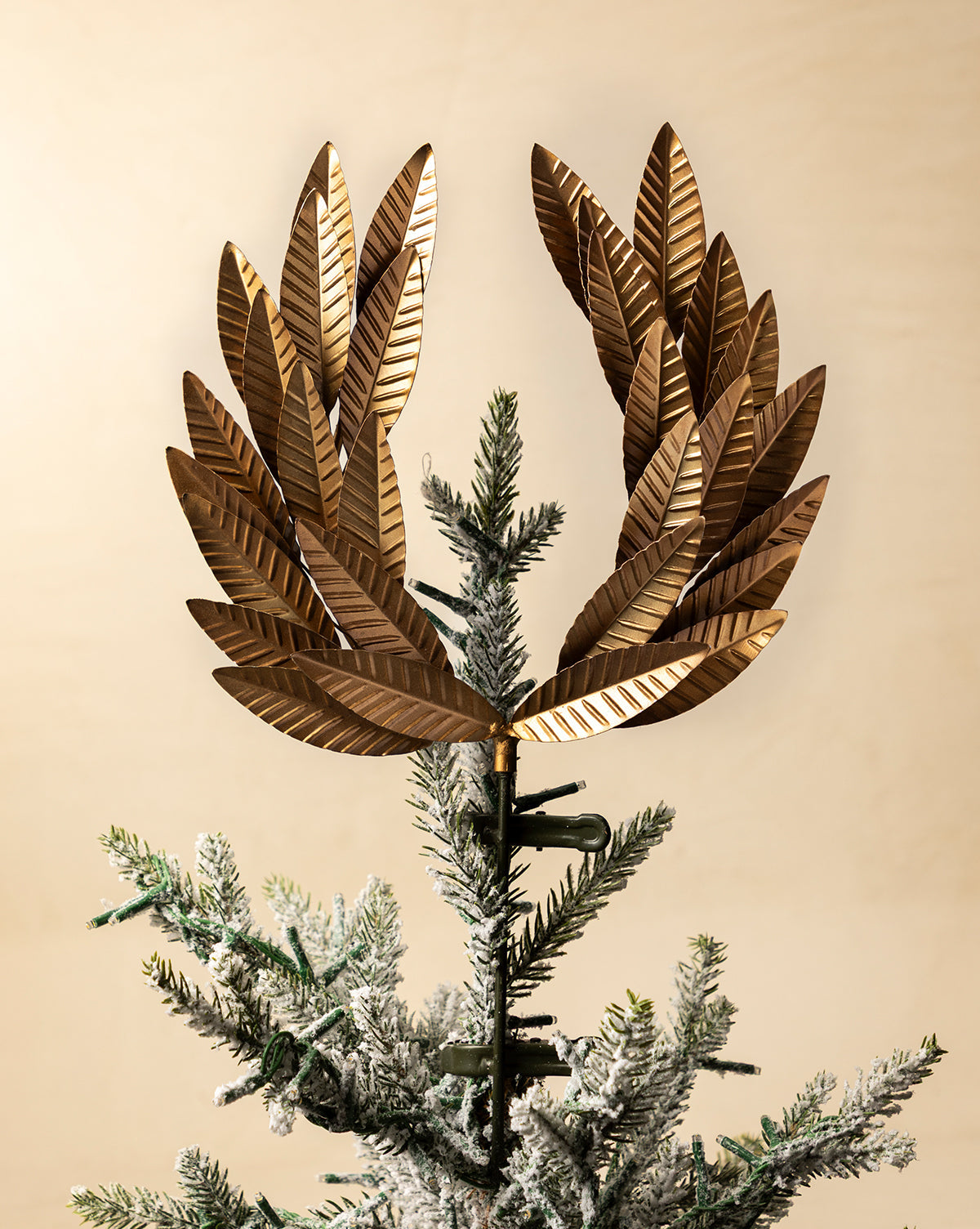 A close-up of a holiday tree features the Laurel Tree Topper in metallic gold, surrounded by green, snow-dusted branches set against a beige background, by McGee & Co.