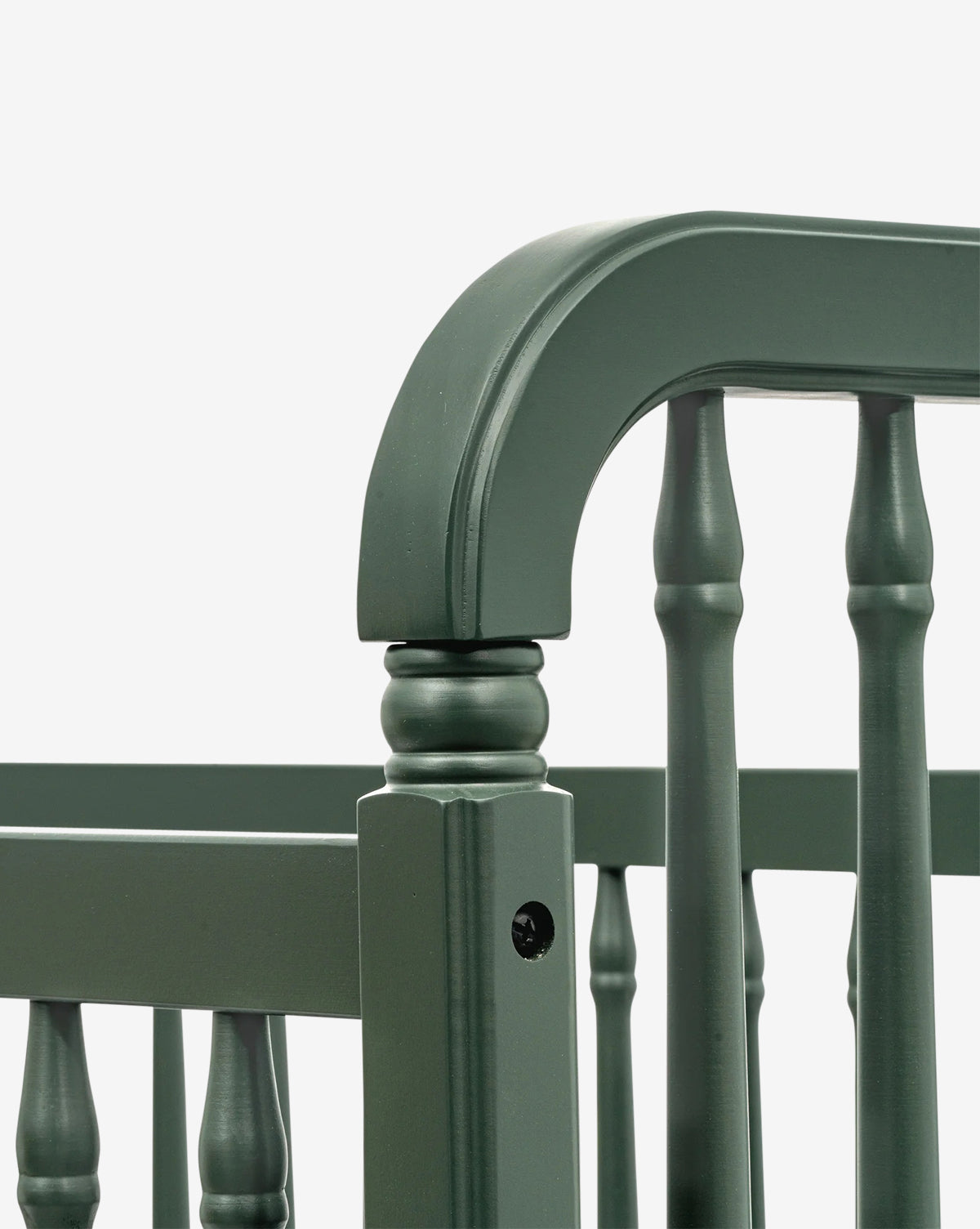 Close-up of the corner of the Liberty 3-in-1 Convertible Spindle Crib with Toddler Bed Conversion Kit in dark green, showing rounded top rails, decorative spindles, and a visible assembly hole on the side panel against a white background by McGee & Co.