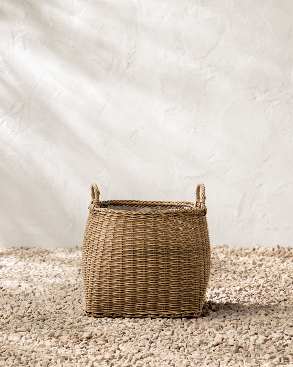 The Lightwash Handled Planter Basket, crafted from woven wicker, rests on small light pebbles against a textured white wall with soft natural light—ideal for showcasing your indoor plants, by McGee & Co.