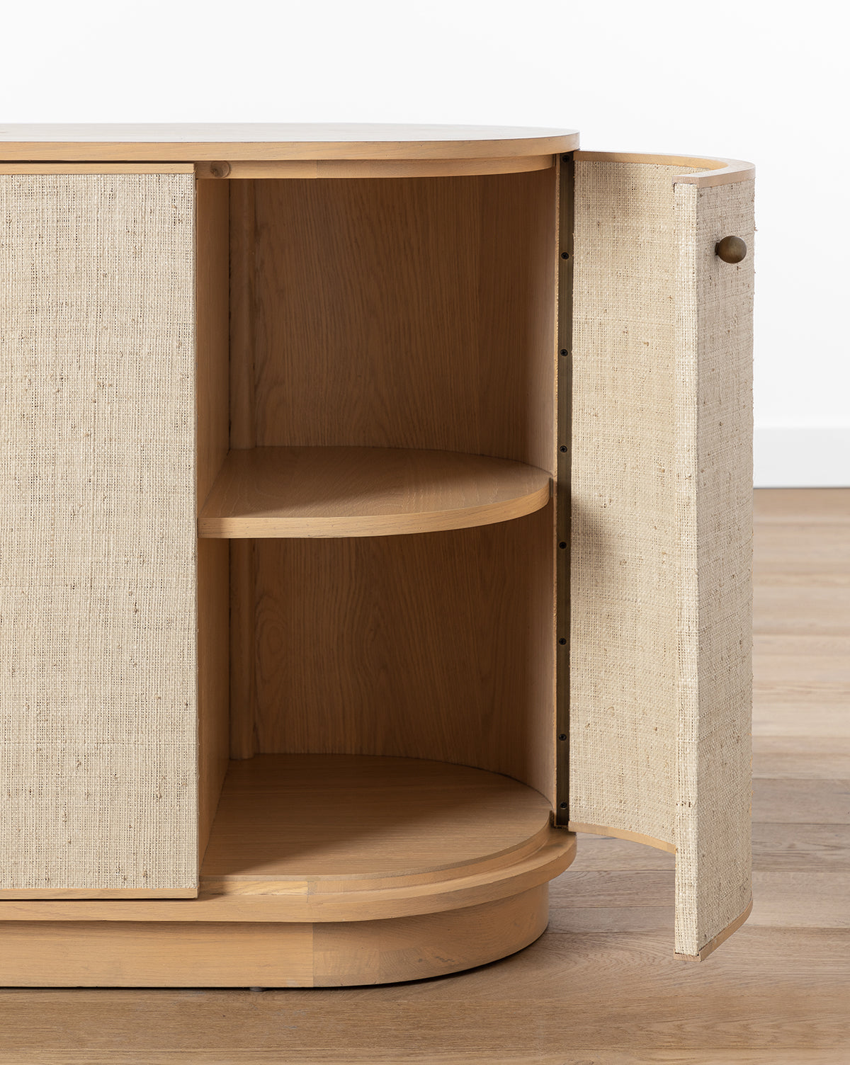 The Linu Woven Sideboard, crafted from white oak, features textured, fabric-like doors—one open to show two curved shelves inside. This modern sideboard stands on a wooden floor against a white wall, by McGee & Co.