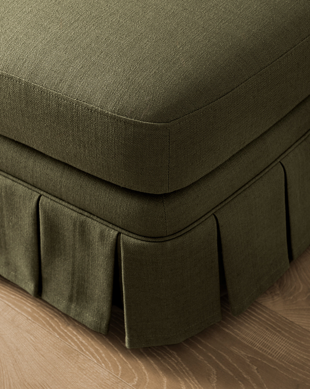 Close-up of the corner of the Louisa Ottoman in olive green upholstery with a box pleat skirt, resting on a light wood floor by McGee & Co.