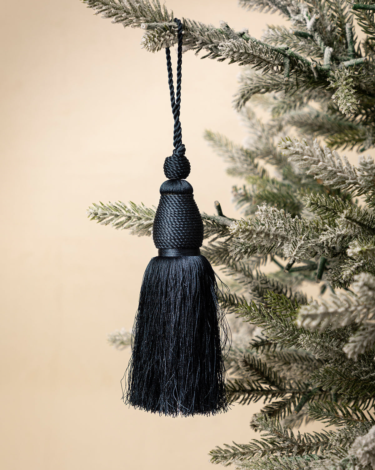A Lucia Bullion Tassel in black brings vintage-inspired charm to your holiday décor, hanging as an ornament from a snow-frosted artificial evergreen branch against a neutral background, by McGee & Co.