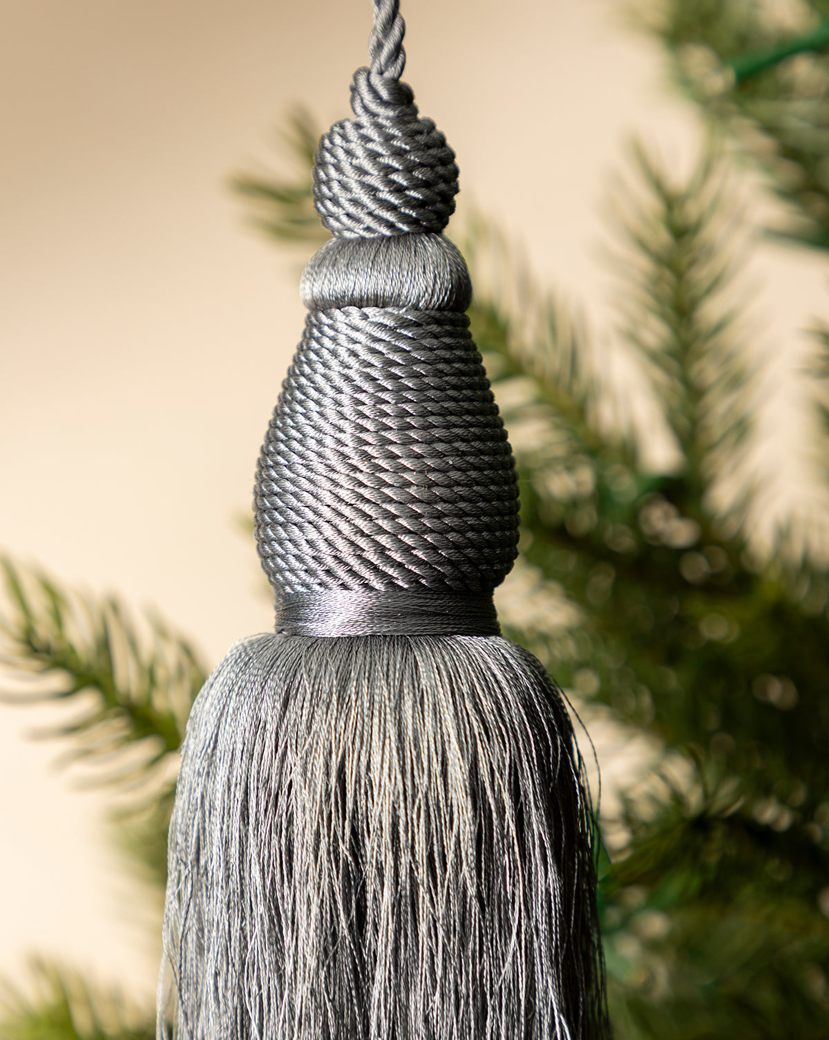 The Lucia Bullion Tassel, a vintage-inspired charm, is shown in close-up against blurred green pine branches and a neutral background, by McGee & Co.