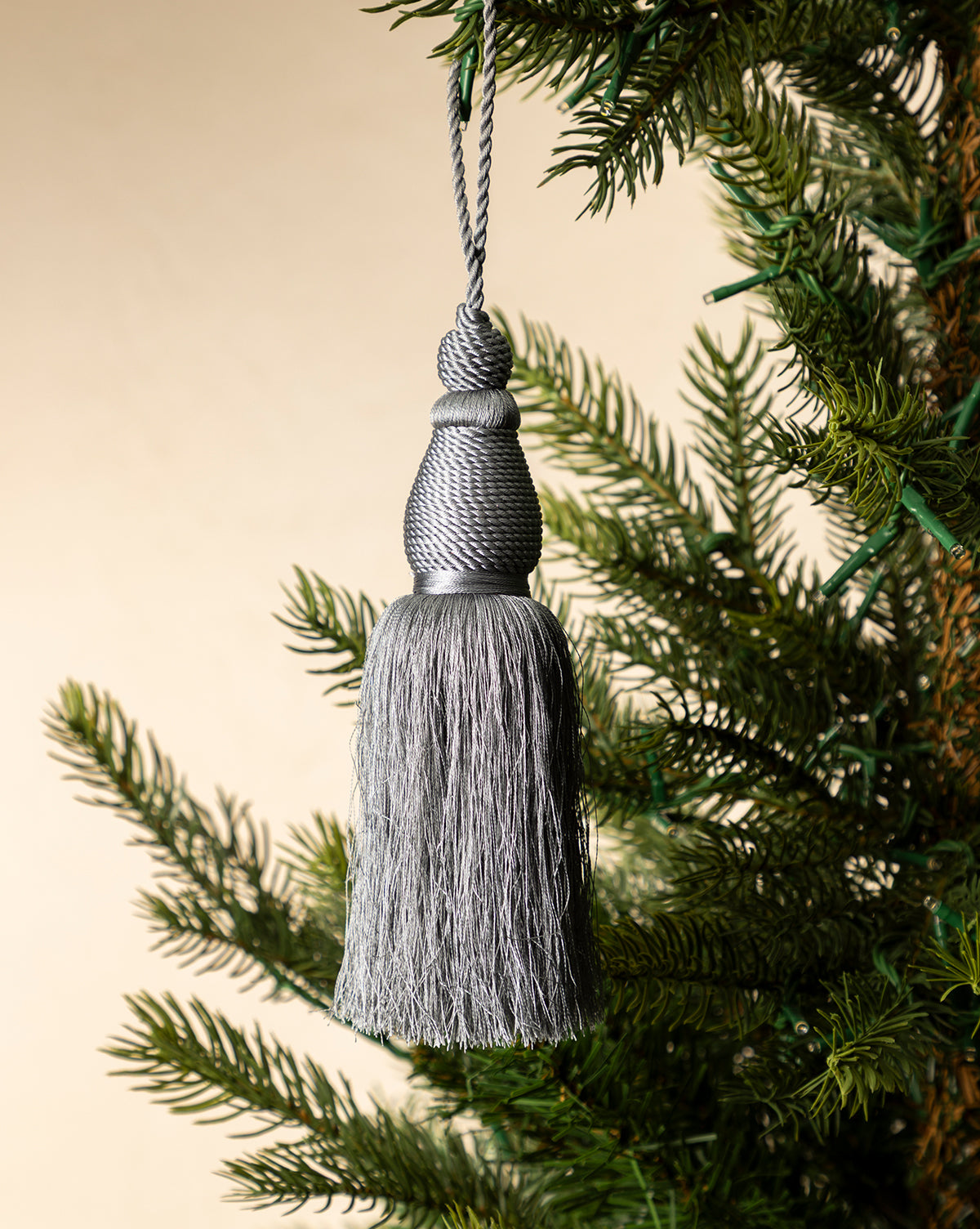 The Lucia Bullion Tassel hangs from a green pine branch, bringing vintage-inspired charm to a neutral beige background, by McGee & Co.