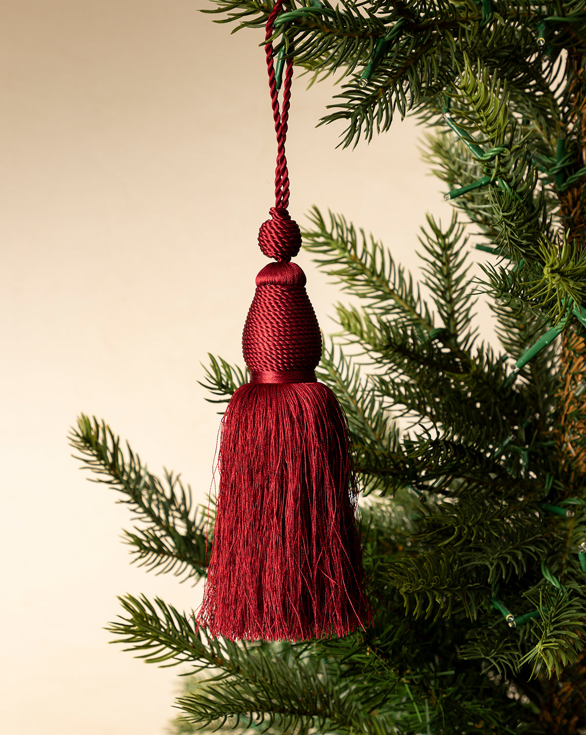 The Lucia Bullion Tassel features deep red hues and vintage-inspired style, hanging from a green pine branch against a soft beige background—perfect for adding charm to your holiday tree ornament display, by McGee & Co.
