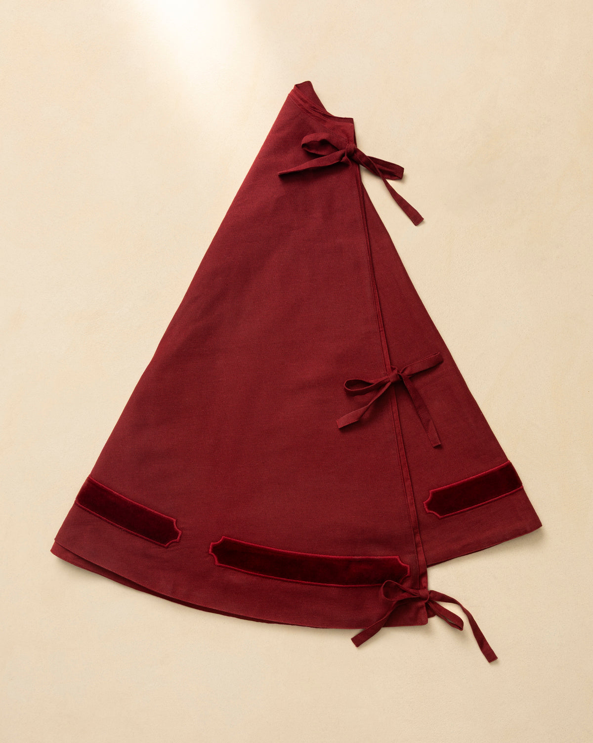 The Mabry Velvet Trimmed Tree Skirt is a deep red, triangular wrap skirt with three side fabric ties and two velvet trims near the hem, shown flat on beige—ideal for adding unique luxury to your holiday decor, by McGee & Co.
