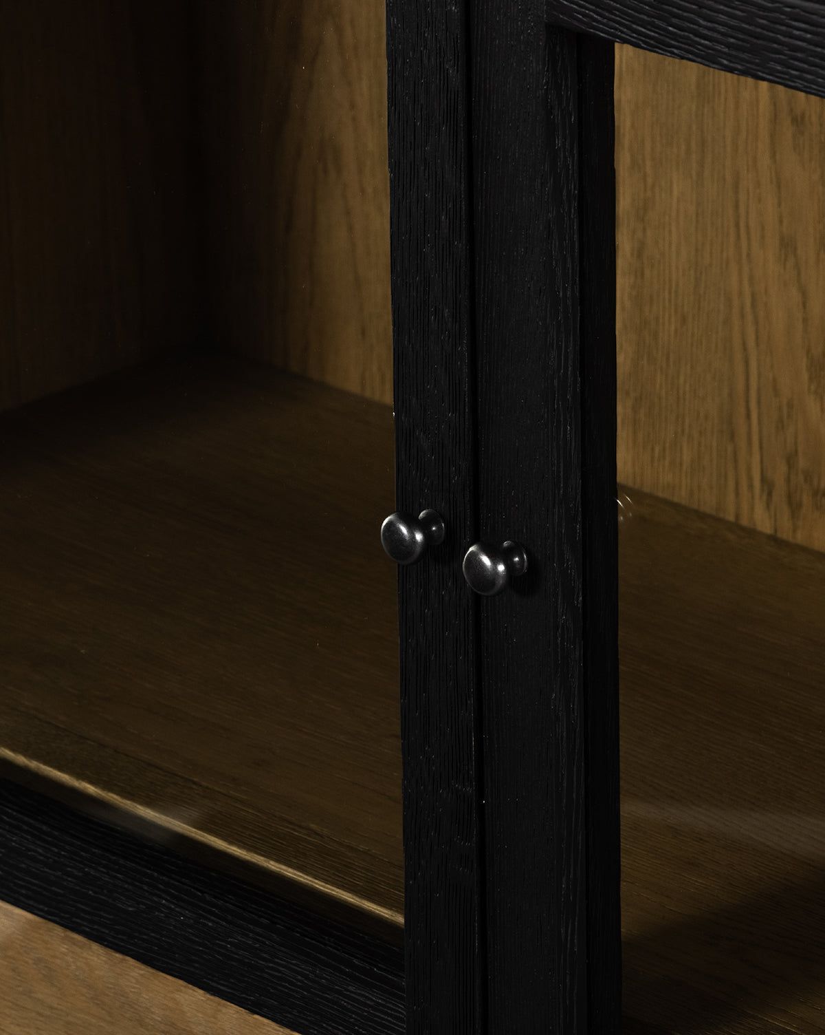Close-up of the Maddox Bookcase’s black-framed glass doors with round metal knobs, showcasing empty wooden shelves. The natural oak finish contrasts with the dark frame for a modern look, by McGee & Co.