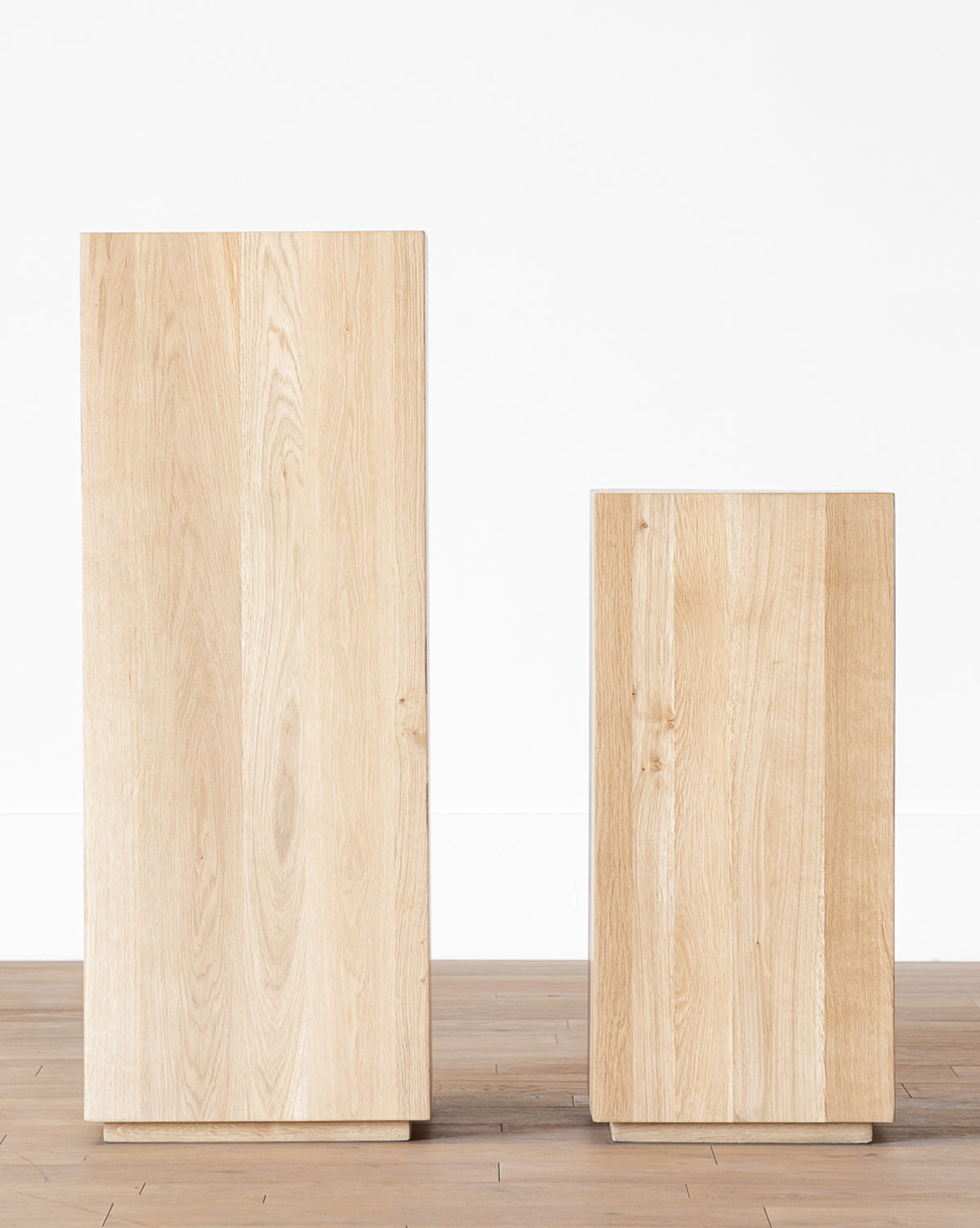 Two rectangular Marlow Pedestal stands of varying heights sit side by side on a wooden floor against a white wall, featuring a light oak finish with visible wood grain for a natural accent, by McGee & Co.