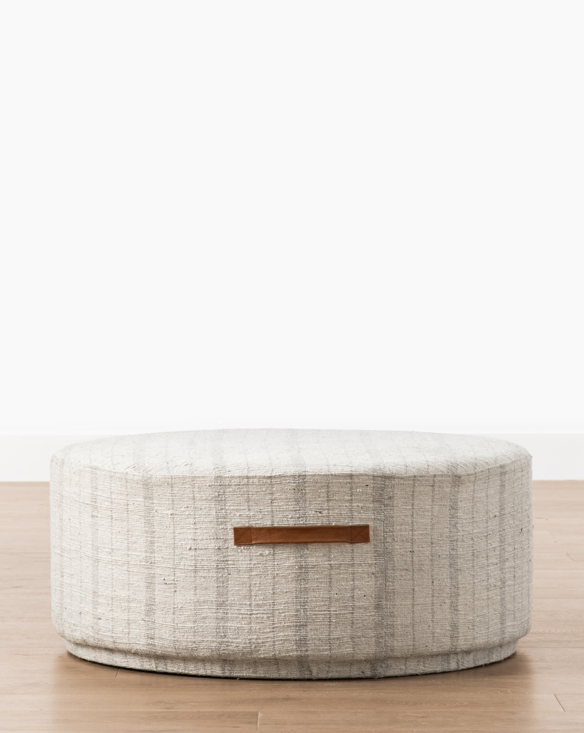 The McKay Round Ottoman features light gray, vertically striped upholstery and a brown leather handle, shown on a wooden floor with a plain white background, by McGee & Co.