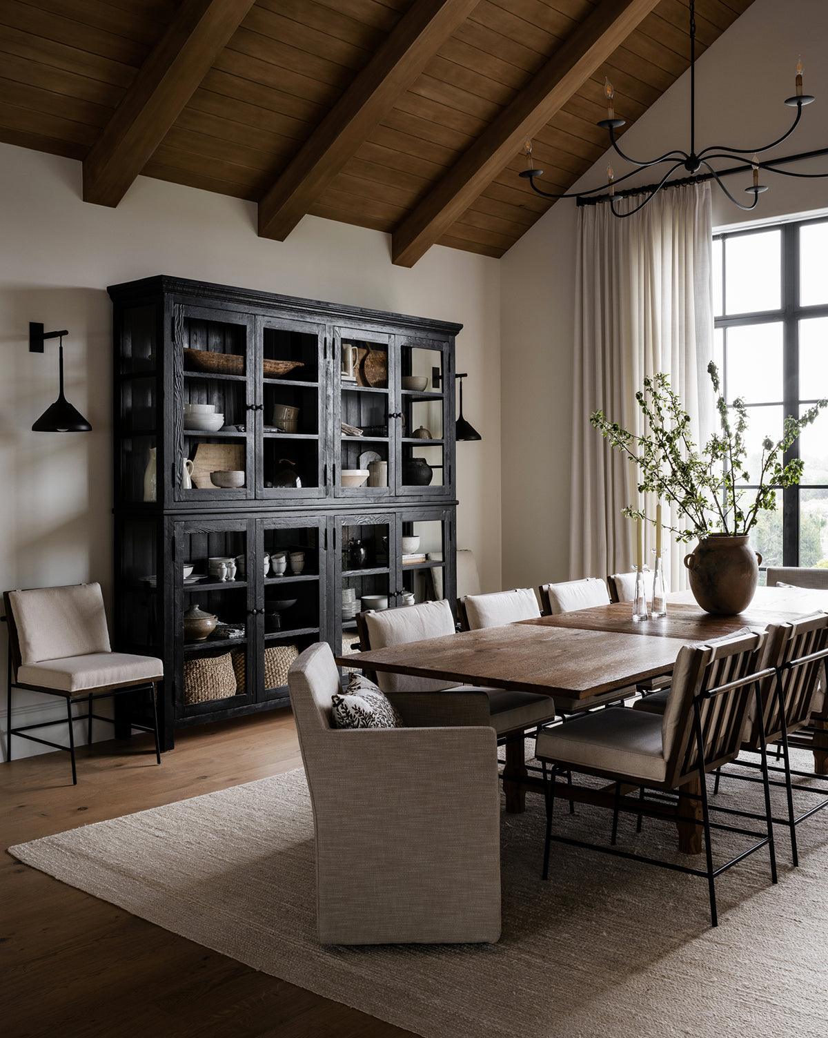 A modern rustic dining room features a large wooden table surrounded by Selas Dining Chairs, a black cabinet with pottery, neutral décor, tall wood-beamed ceilings, a light rug, and windows with sheer curtains by McGee & Co.