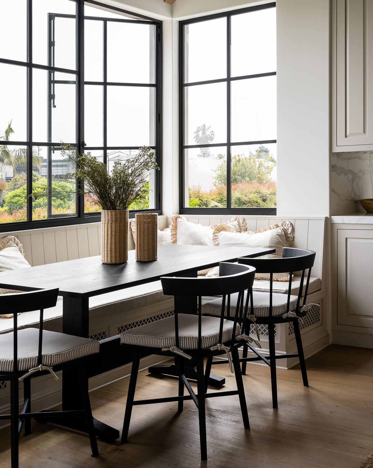 A modern dining area with a black table, Reeves Dining Chair, a built-in bench with cushions, large windows for natural light, and decorative vases filled with greenery on the table, by McGee & Co.