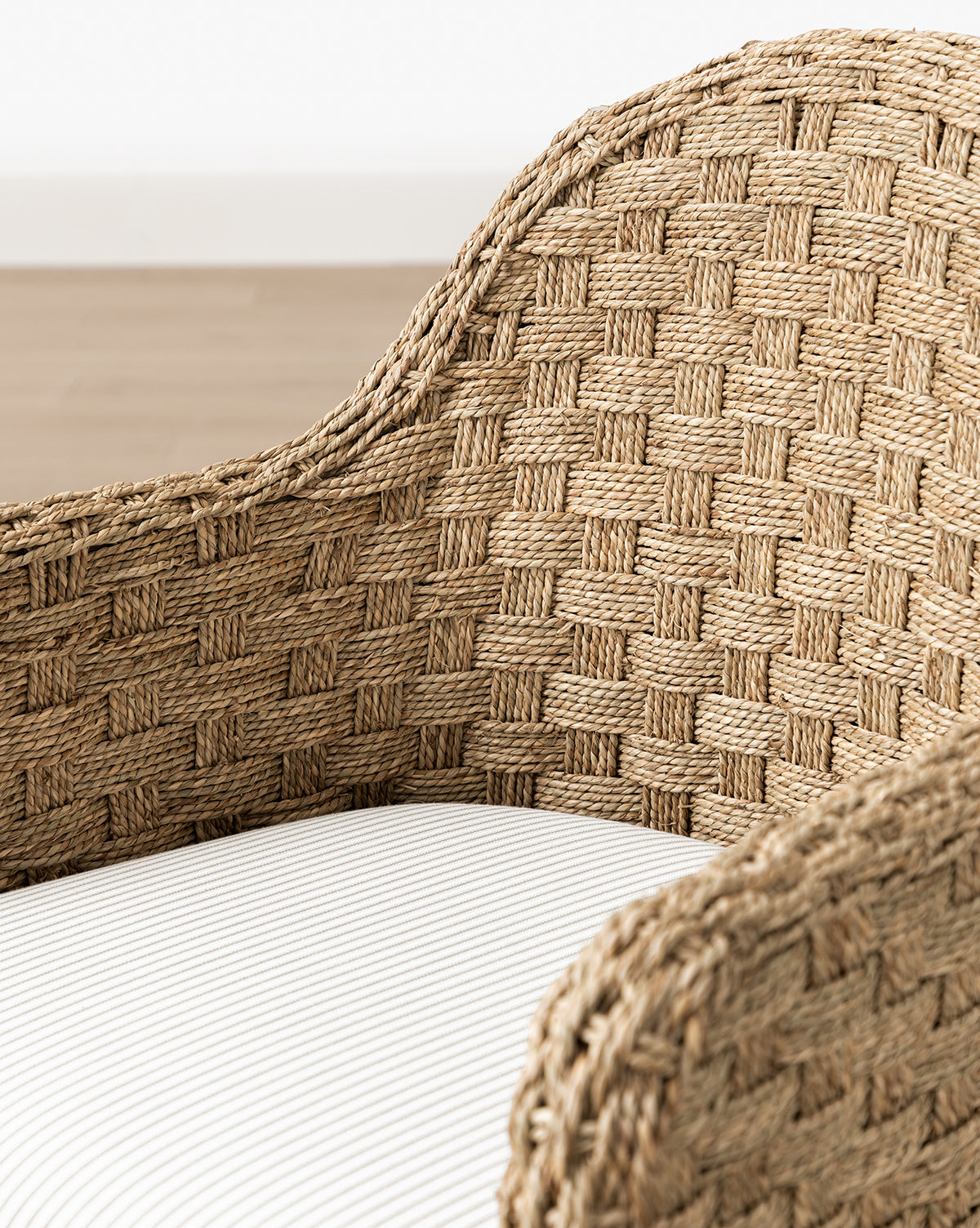 Close-up of the Molly Arm Chair, a modern design with a curved back and armrest, showcasing basketweave detailing and a white striped seat cushion. A blurred wooden floor and white wall appear in the background, by McGee & Co.