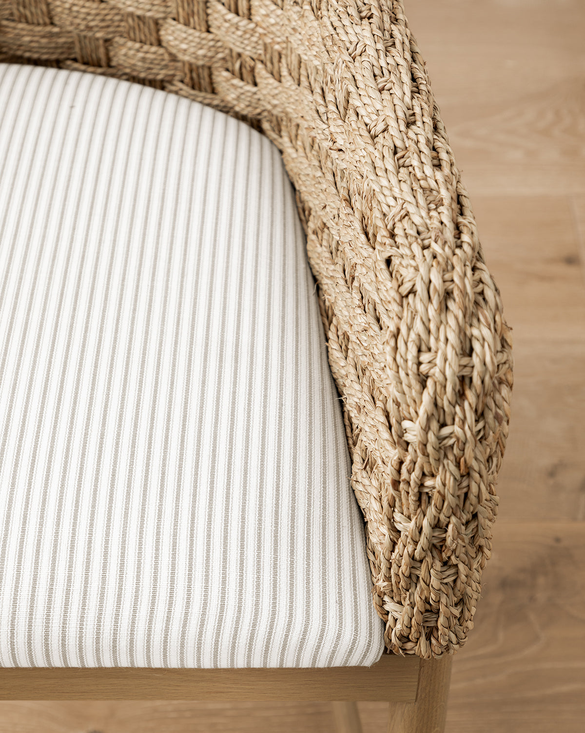 Close-up of the Molly Arm Chair, a modern design featuring a woven rattan frame, curved armrests, and a white seat cushion with thin light gray vertical stripes, shown resting on a wooden floor, by McGee & Co.