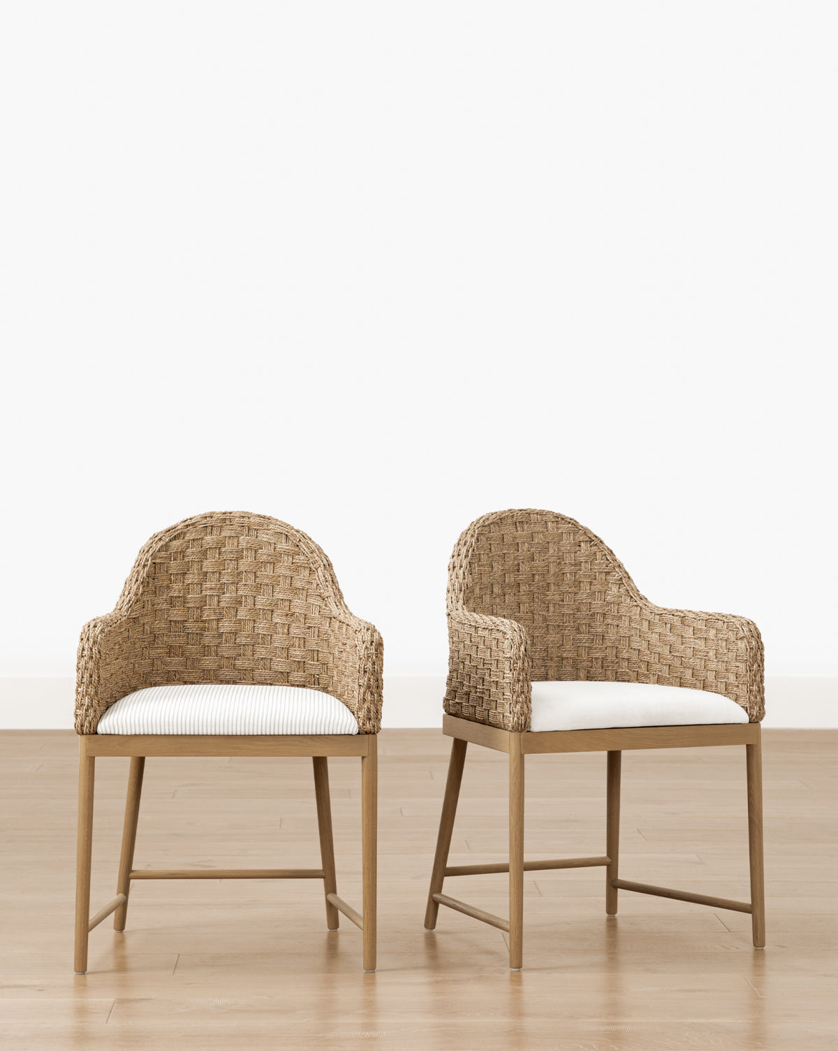 Two Molly Arm Chairs with curved wicker backs and white seat cushions are positioned side by side on a light wood floor against a plain white wall by McGee & Co.