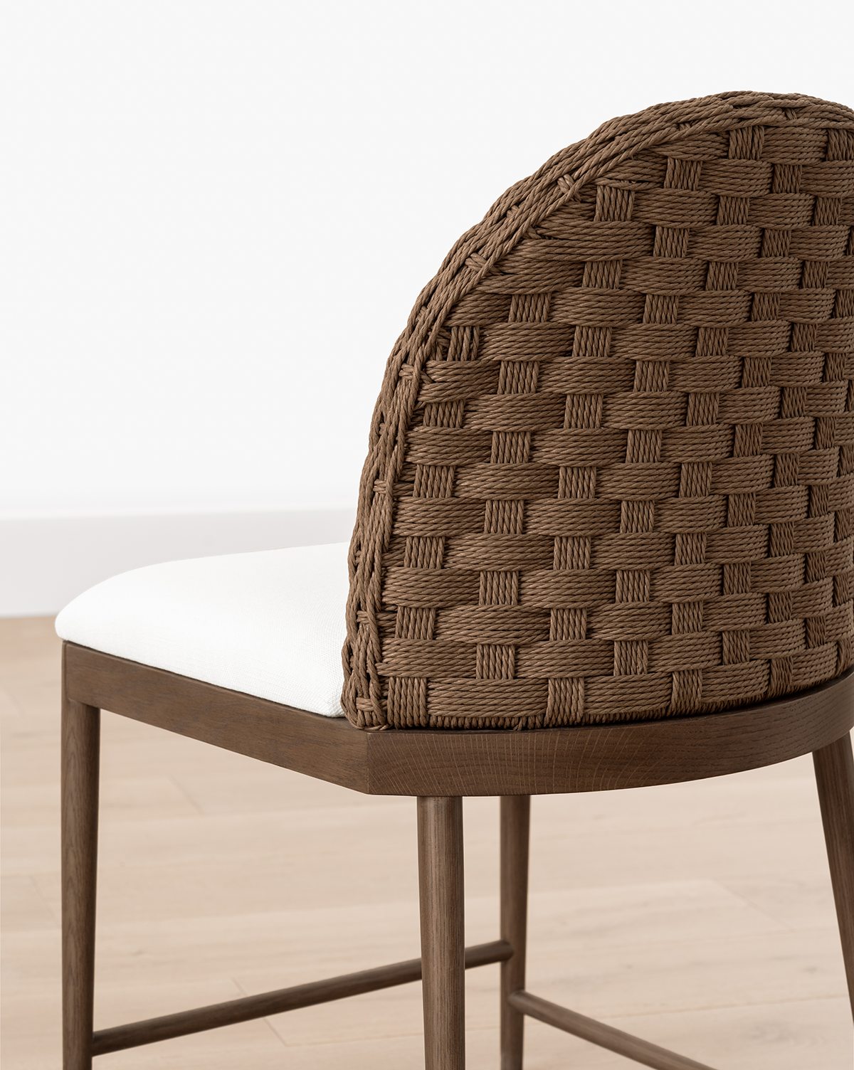 The Molly Dining Chair features a woven brown backrest, white cushioned seat, and sturdy wooden legs, shown here on a light wood floor against a white wall, by McGee & Co.