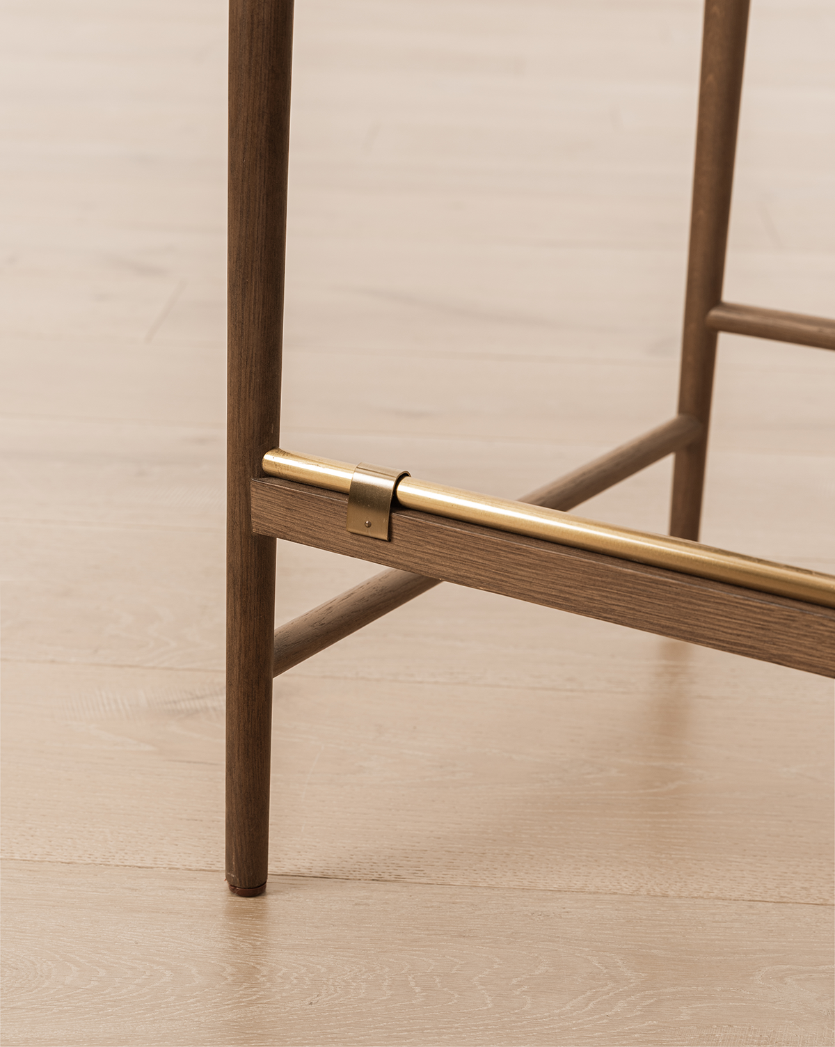 Close-up of the Molly Stool’s solid oak leg with a brass footrest near the base on a light wood floor—perfect refined detailing for this natural bar or counter stool, by McGee & Co.