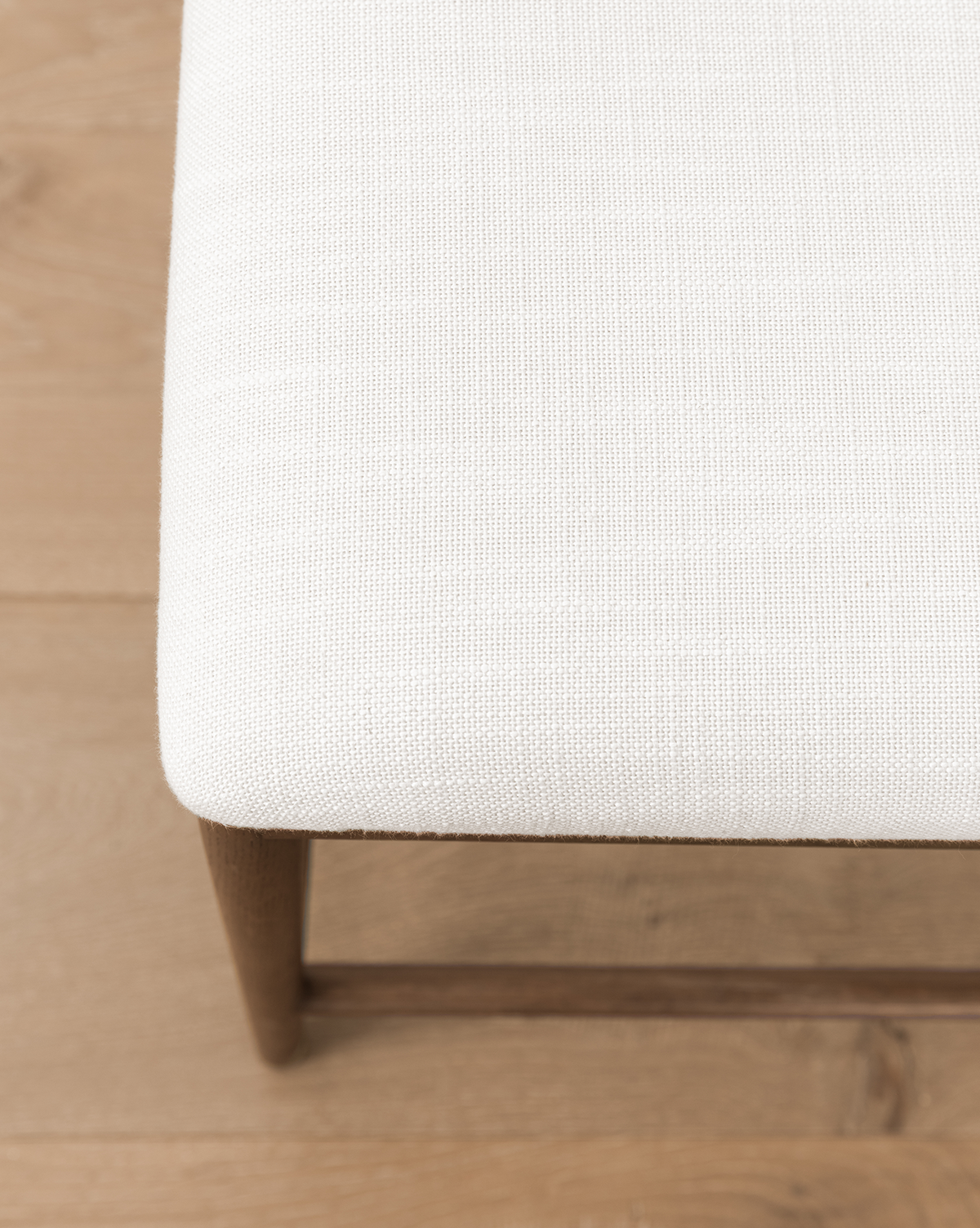 A detailed view of the Molly Stool’s corner highlights its solid oak frame and textured off-white fabric seat, set atop a light wooden floor, by McGee & Co.