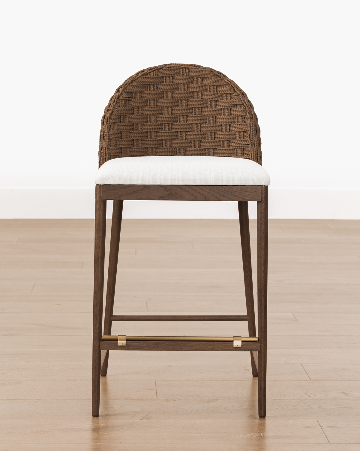 The Molly Stool features a woven brown backrest, white cushioned seat, and solid oak legs with a brass footrest, shown on a light wooden floor against a white wall, by McGee & Co.