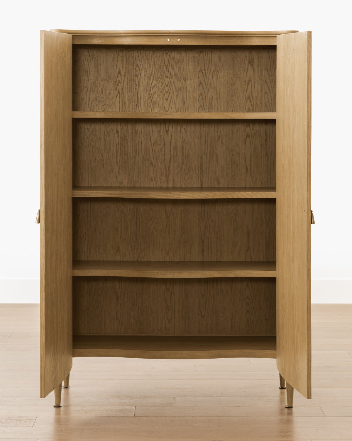 The Moorland Cabinet, with two open doors, reveals three spacious shelves inside—ideal for living room storage. Standing on four legs, its set on a light wooden floor against a plain white wall by McGee & Co.