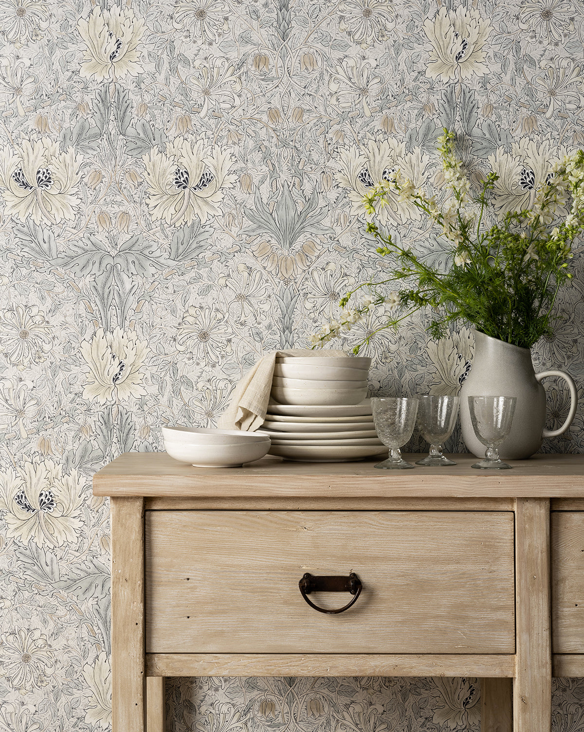 A rustic wooden side table with a drawer holds stacked white dishes, cloth napkins, clear glasses, and the Levi Stoneware Pitcher filled with green and white flowers. Muted floral-patterned wallpaper sets the background by McGee & Co.