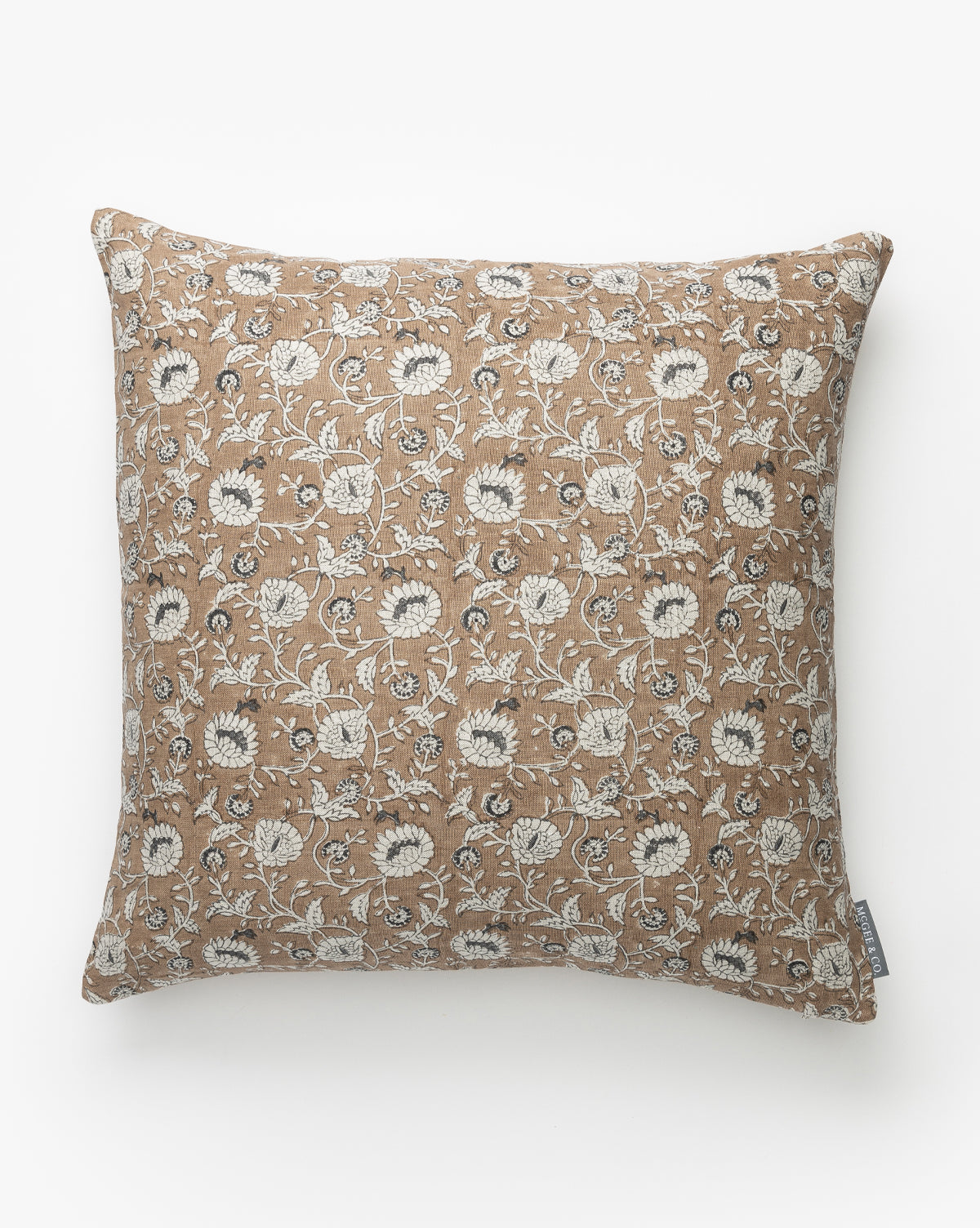 The Nanette Pillow Cover features a square shape with a light brown background and white floral patterns of leaves and flowers. It has a small fabric tag on one side and is displayed on a plain white surface by McGee & Co.