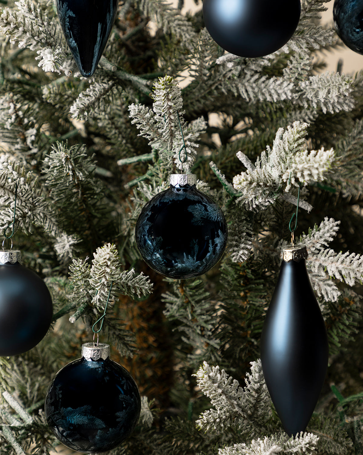 Close-up of a holiday tree decorated with Assorted Ornaments Navy (Set of 10) in shiny dark blue and black, round and elongated shapes, hanging from green branches dusted with artificial snow, by McGee & Co.