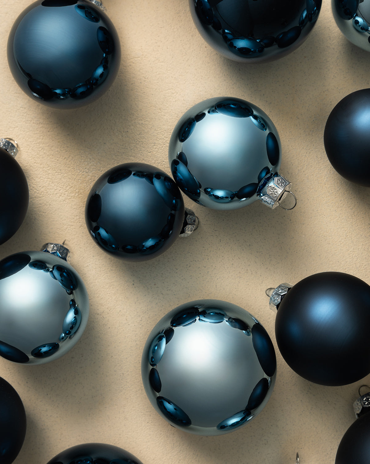 Navy Glass Baubles (Set of 21 Assorted) with shiny and matte finishes, arranged on a beige surface, reflect light and create a festive holiday display, by McGee & Co.