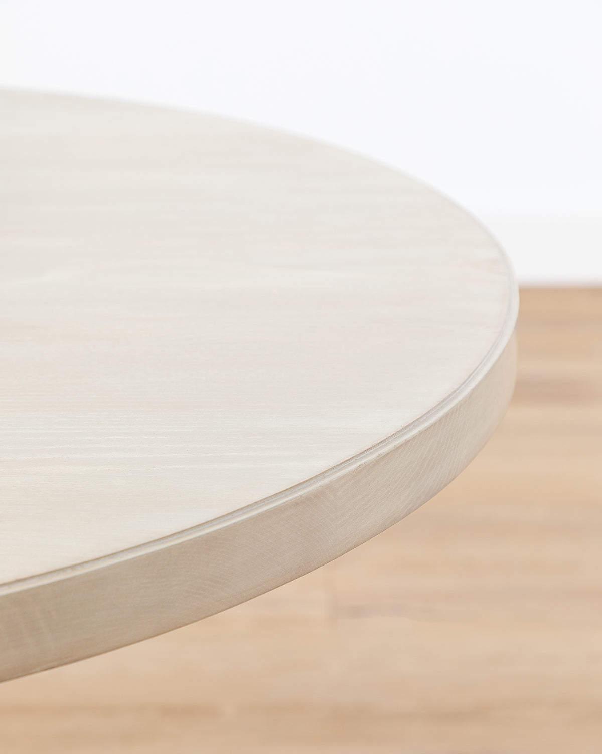 A close-up of the smooth, light-toned edge of the round Orla Dining Table is set against a blurred rustic oak floor and a white background, by McGee & Co.