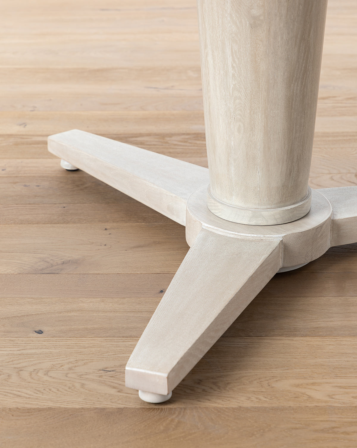 Close-up of the Orla Dining Table’s base in a light finish, showing its central cylindrical support and three wide, angled legs on a light brown wooden floor, by McGee & Co.