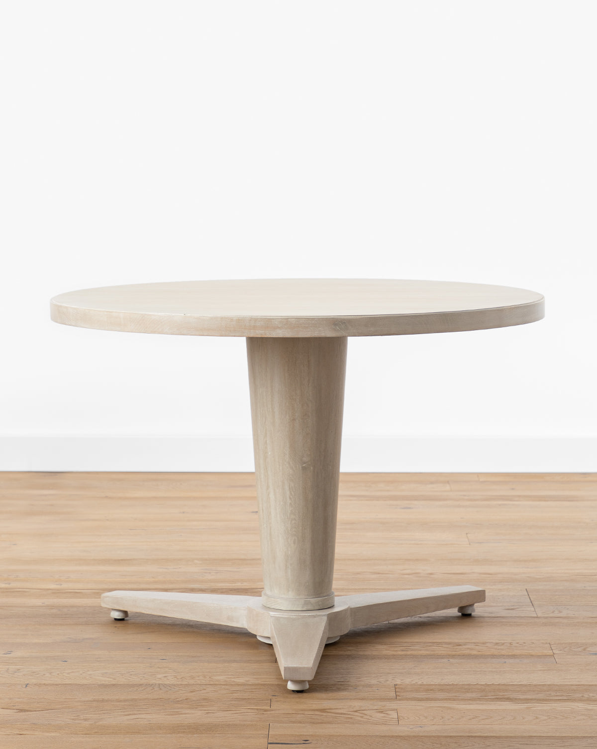 The Orla Dining Table showcases a round rustic oak top, a sturdy pedestal base with three feet, and is displayed on a light wood floor with a plain white background by McGee & Co.