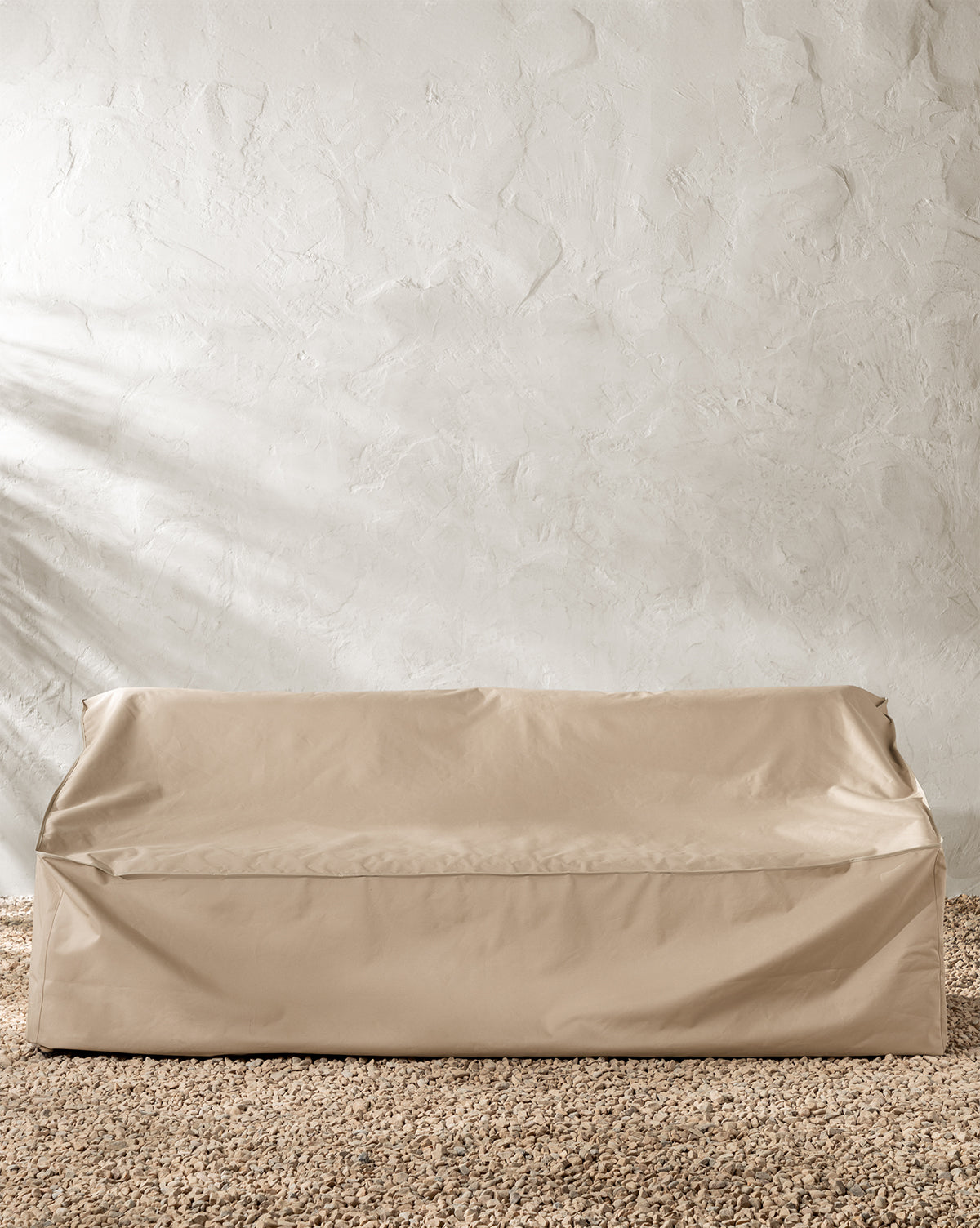 A beige, rectangular outdoor sofa protected by the Outdoor Sofa Cover rests on gravel against a textured off-white wall, by McGee & Co.