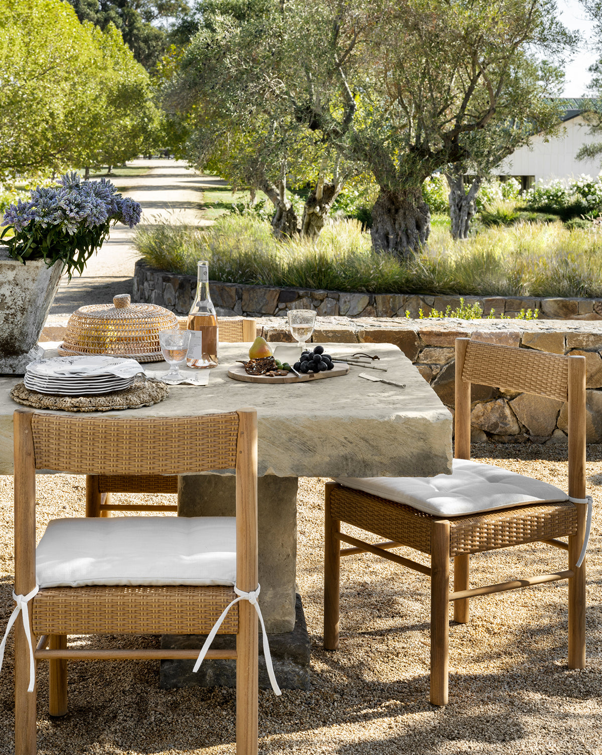 A rustic outdoor dining area with a stone table set for wine, fruit, and cheese features Simeon Outdoor Dining Chairs, surrounded by lush greenery, olive trees, and fragrant lavender, by McGee & Co.