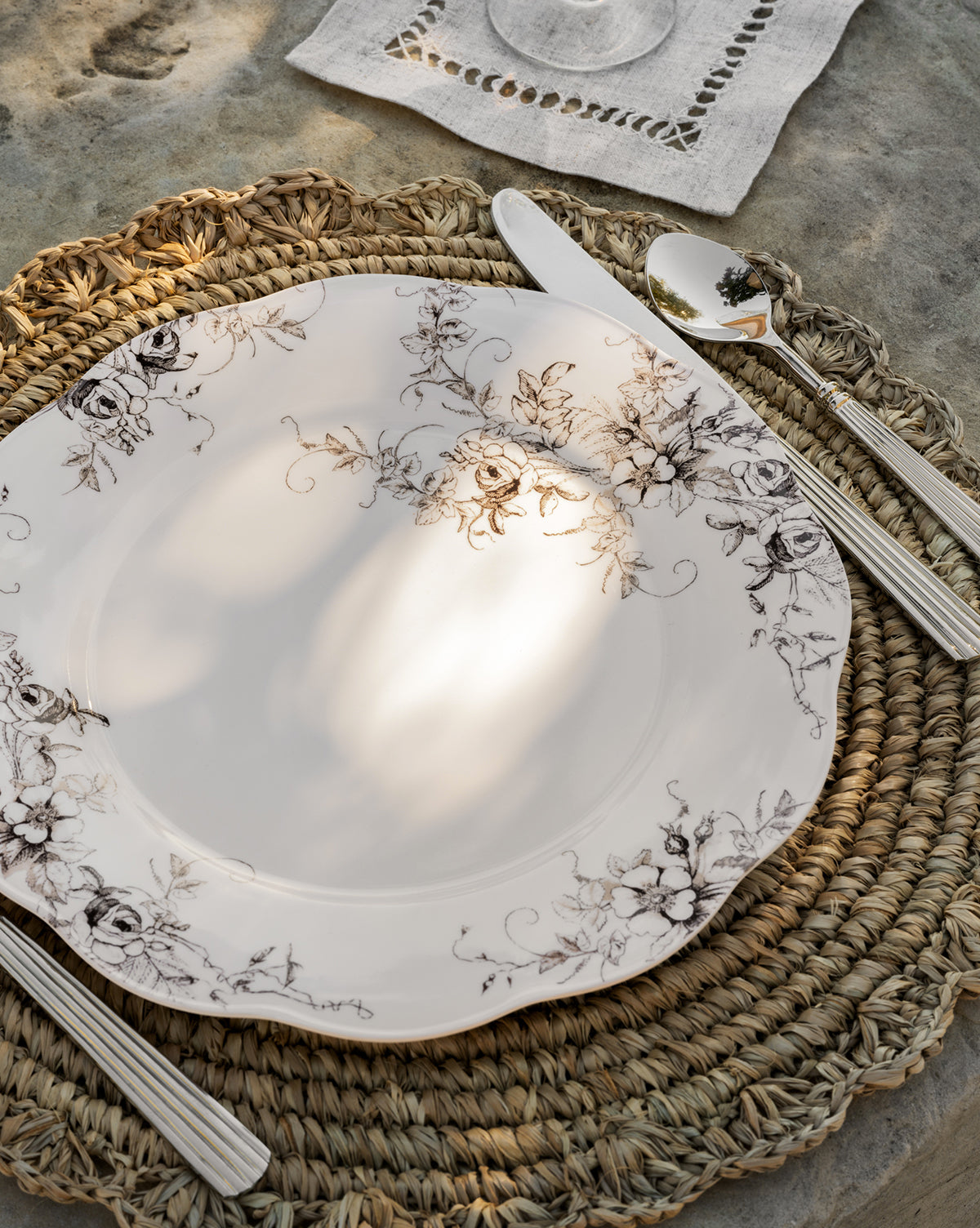 An Avondale Melamine Dinner Plate in white with a black floral motif sits on a woven placemat with cutlery, a linen napkin, and a glass atop a textured stone surface as sunlight casts gentle shadows. Set includes 4 plates by McGee & Co.