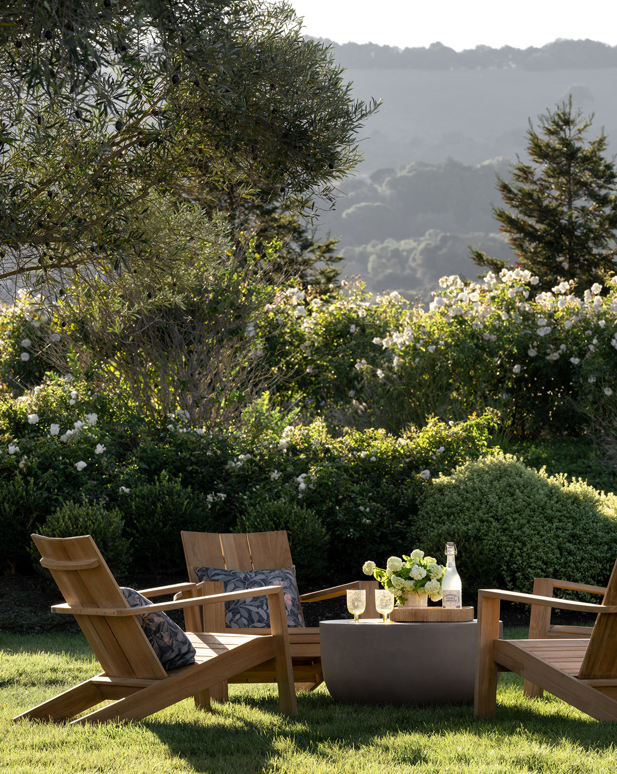 Two Triby Teak Outdoor Lounge Chairs with cushions sit by a small patio table with wine, glasses, and flowers in a lush garden with blooming bushes and distant hills—ideal for stylish all-weather relaxation, by McGee & Co.