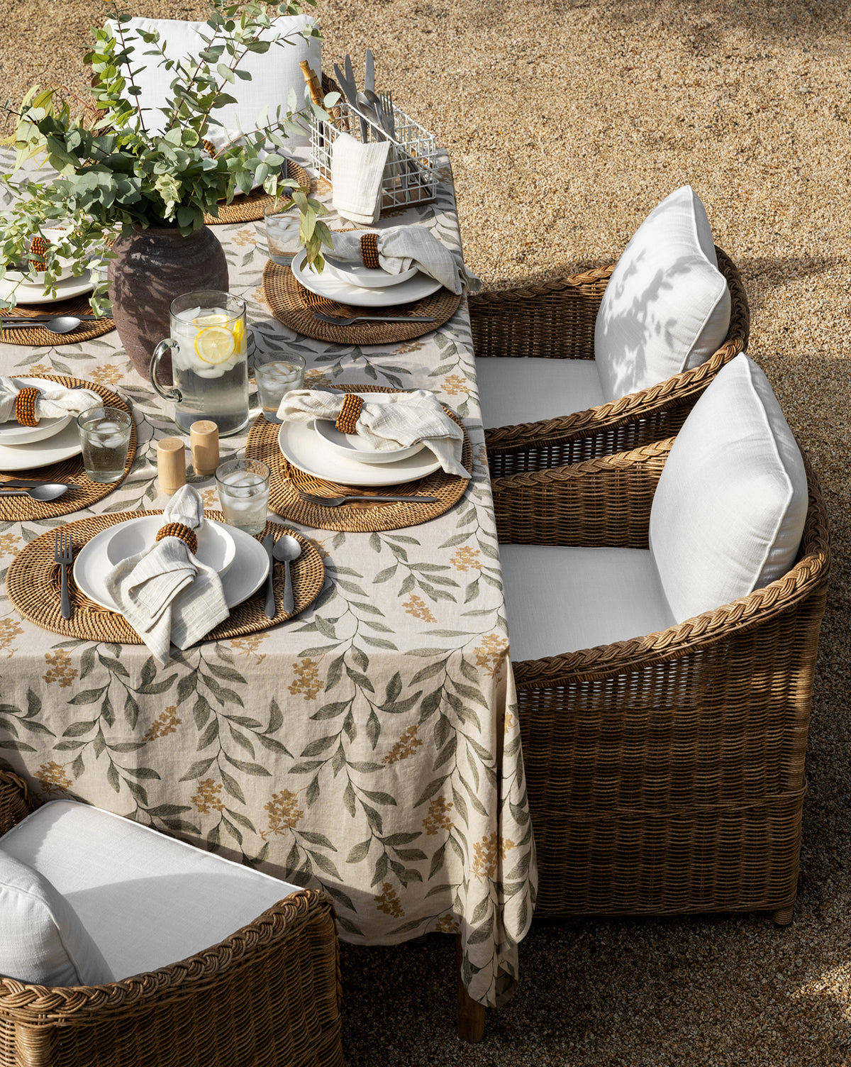 A rectangular outdoor dining table set for six features woven placemats, white plates, napkins with rings, a vase with greenery, and Haviland Outdoor Dining Chairs with white cushions on a gravel surface by McGee & Co.