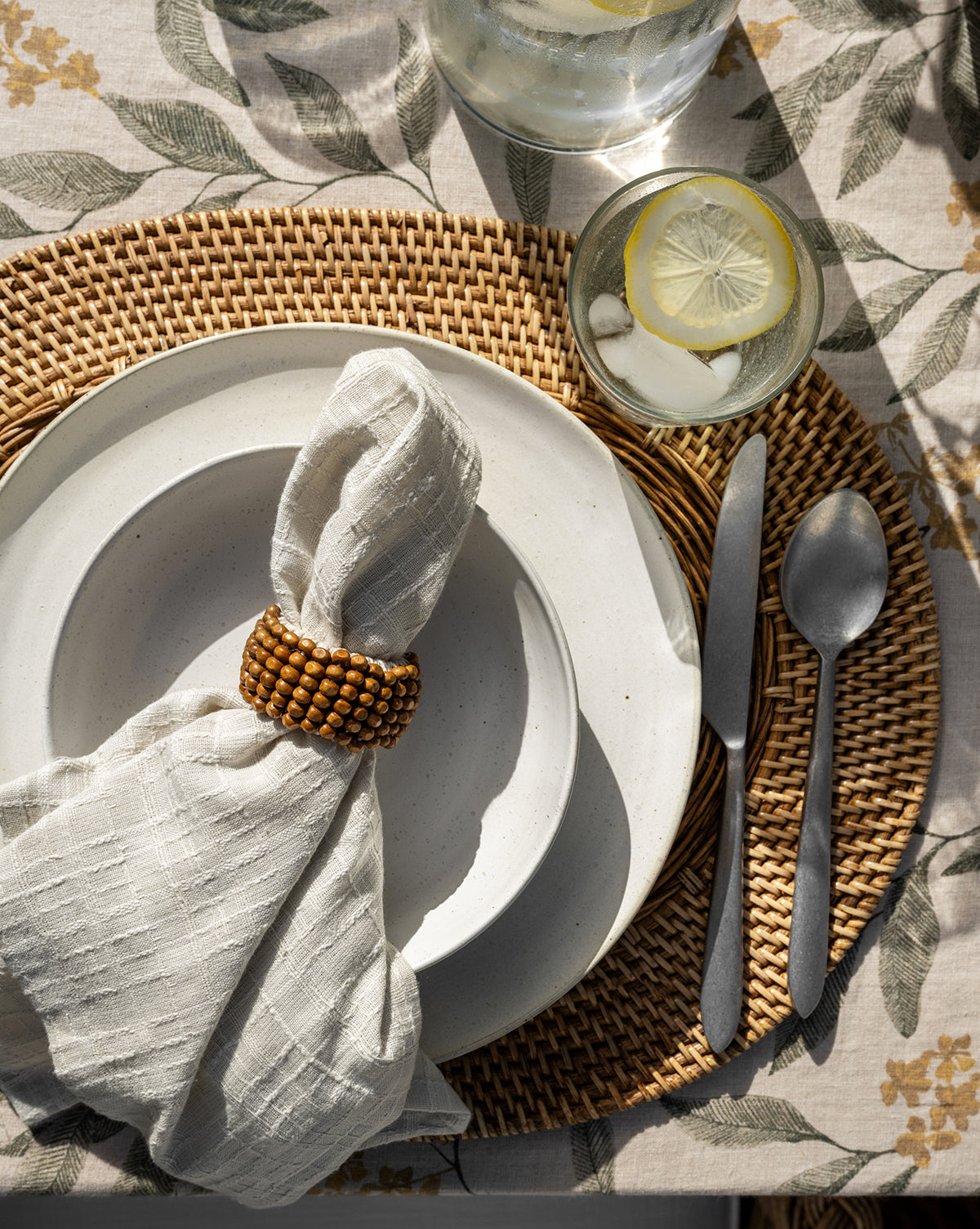 A table setting features a white plate, a cloth napkin in a beaded ring, the Lilia Flatware (Set of 5) on a woven placemat, and a glass of ice water with lemon, all arranged on a floral tablecloth.