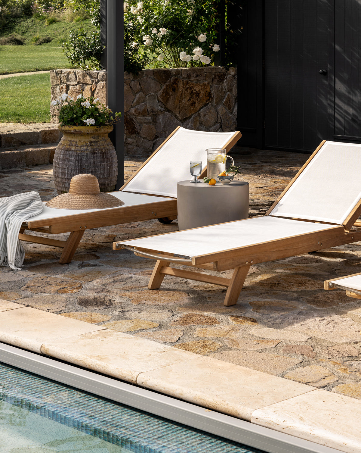 Two Cadie Outdoor Chaise lounge chairs with white, weather-resistant cushions sit poolside on a stone patio. A small table holds drinks and a pitcher, while a straw hat and striped towel rest on one chair amid lush greenery, by McGee & Co.