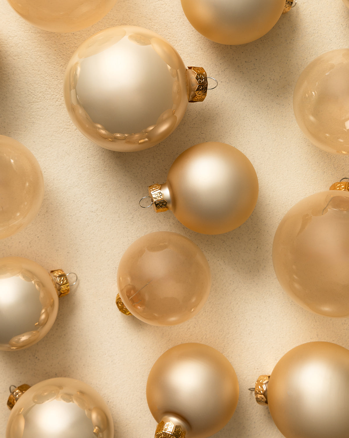 The Pearl Glass Baubles (Set of 21 Assorted) features gold, champagne, and pearl ornaments with metallic caps, beautifully arranged on a beige textured surface for a festive and elegant display in soft neutral tones, by McGee & Co.
