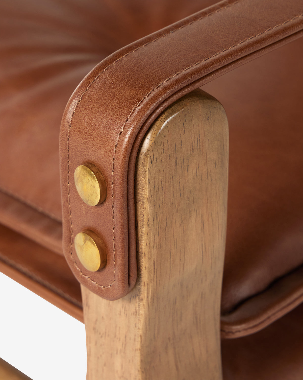 Close-up of the Pederson Dining Armchair armrest, highlighting its brown leather upholstery, brass buttons, and a smooth, honey-finished wood frame with a top grain leather seat, by McGee & Co.