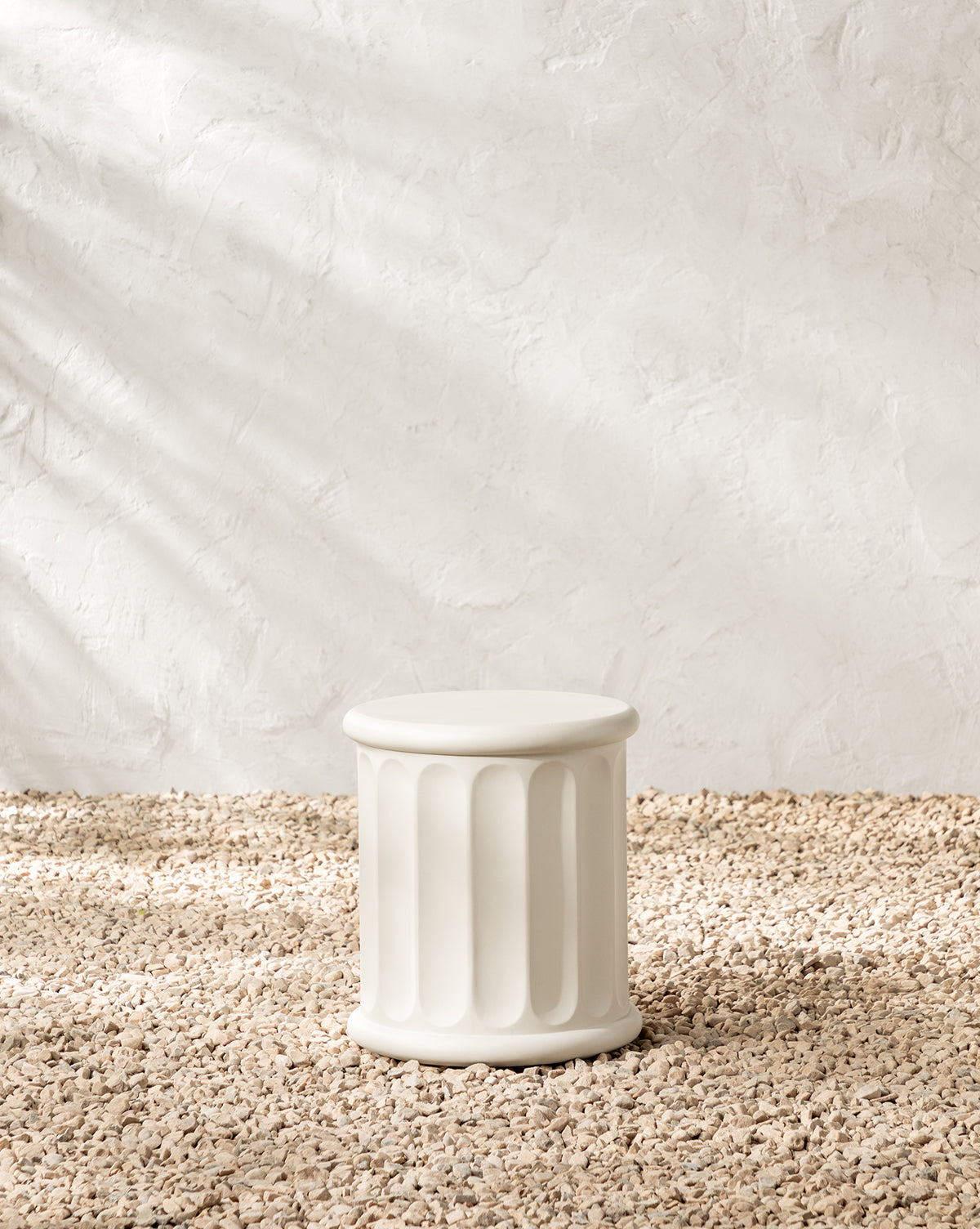 The Penelope Outdoor Side Table, a white modern piece made from fiber cement with vertical grooves, stands on a pebbled surface against a textured light wall in soft natural lighting, by McGee & Co.